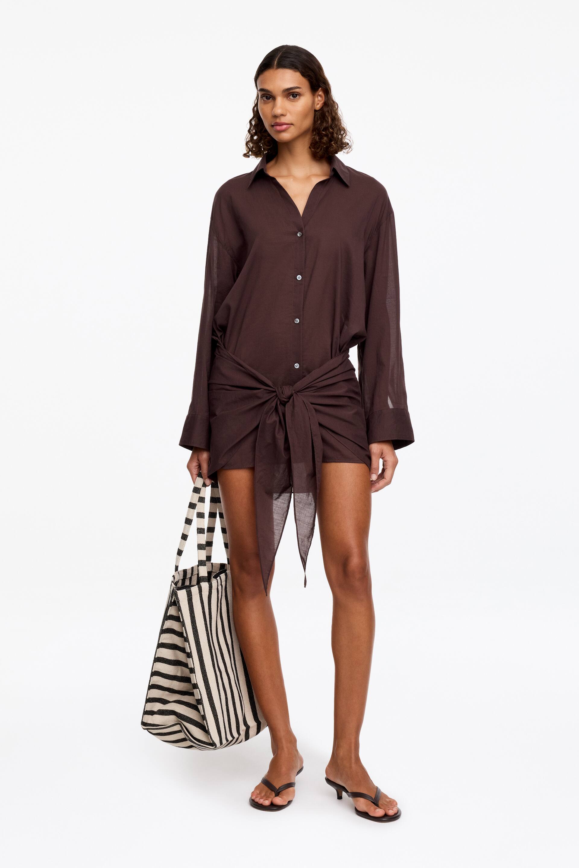Visualizza immagine più grande: La modella indossa un abito camicia corto marrone con bottoni lungo tutta la parte anteriore e maniche lunghe con polsini. L'abito presenta un dettaglio con nodo legato in vita sul davanti.