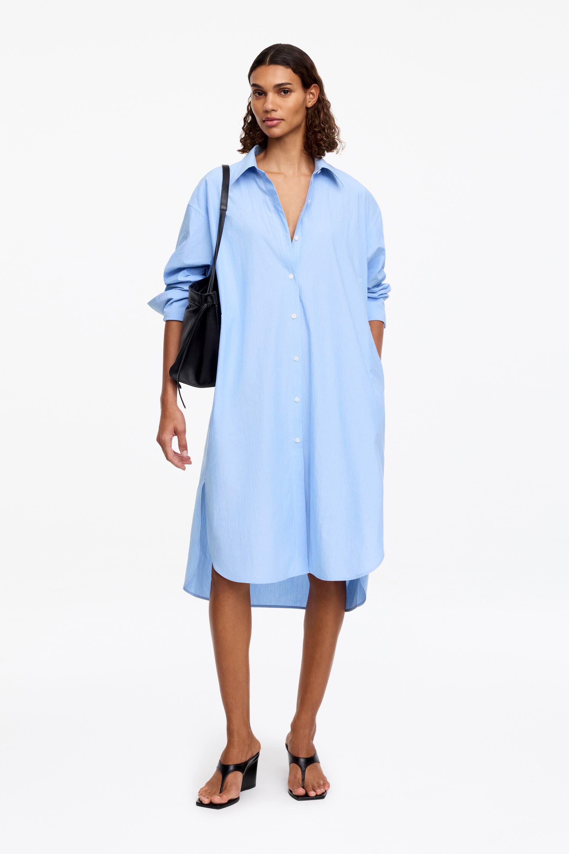 Größeres Bild ansehen: Ein hellblaues, locker sitzendes Hemdkleid mit einem subtilen Streifenmuster wird von dem Model getragen. Es verfügt über einen klassischen Kragen, lange Ärmel mit hochgeschobenen Manschetten und Seitentaschen. Eine Hand des Models ruht in einer Tasche, und eine schwarze Schultertasche wird über dem knielangen Kleid getragen, das einen abgerundeten Saum mit einem längeren Rückenteil hat.