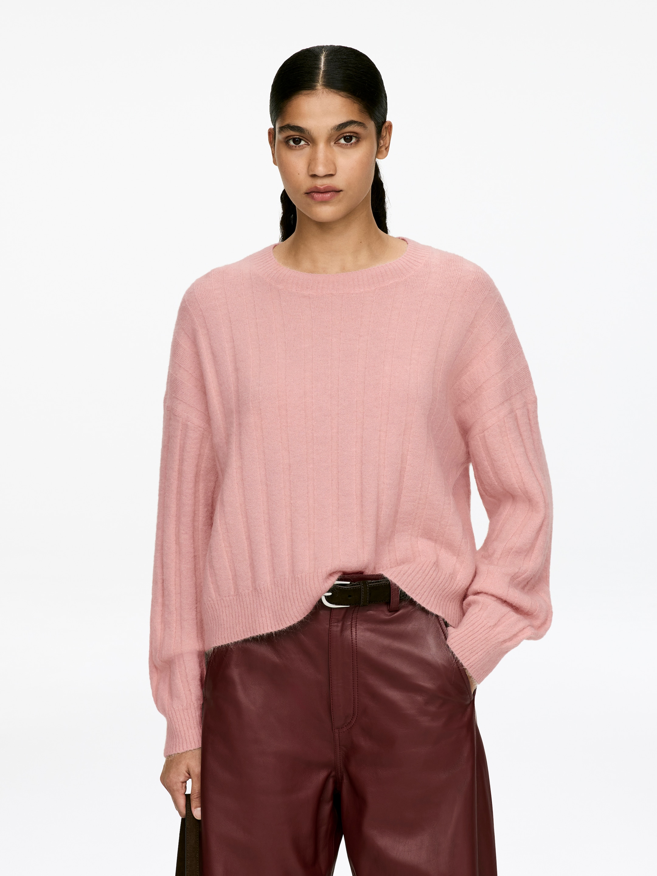 Grösseres Bild ansehen: Ein rosa, breit geripptes Pullover mit Rundhalsausschnitt und langen Ärmeln wird von dem Modell getragen. Der locker sitzende Pullover fällt über die Taille des Modells und das Modell steckt eine Hand in eine Tasche, um den Saum des Kleidungsstücks zu präsentieren.