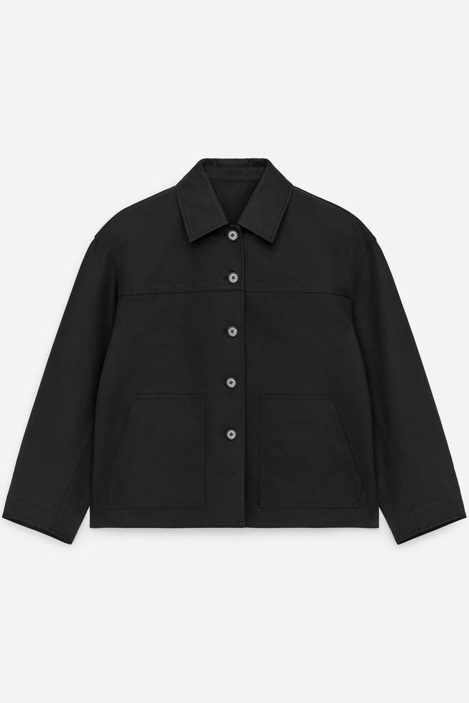 Eine schwarze Overshirt ist nach VORNE gerichtet und präsentiert eine subtil verkürzte, von Arbeitskleidung inspirierte Silhouette aus einem robusten Stoff. Es verfügt über einen spitzen Kragen, einen Knopfverschluss vorne mit vier sichtbaren Knöpfen und zwei großen aufgesetzten Taschen an der unteren Vorderseite, mit einer horizontalen Naht, die über die Brust verläuft. - 1