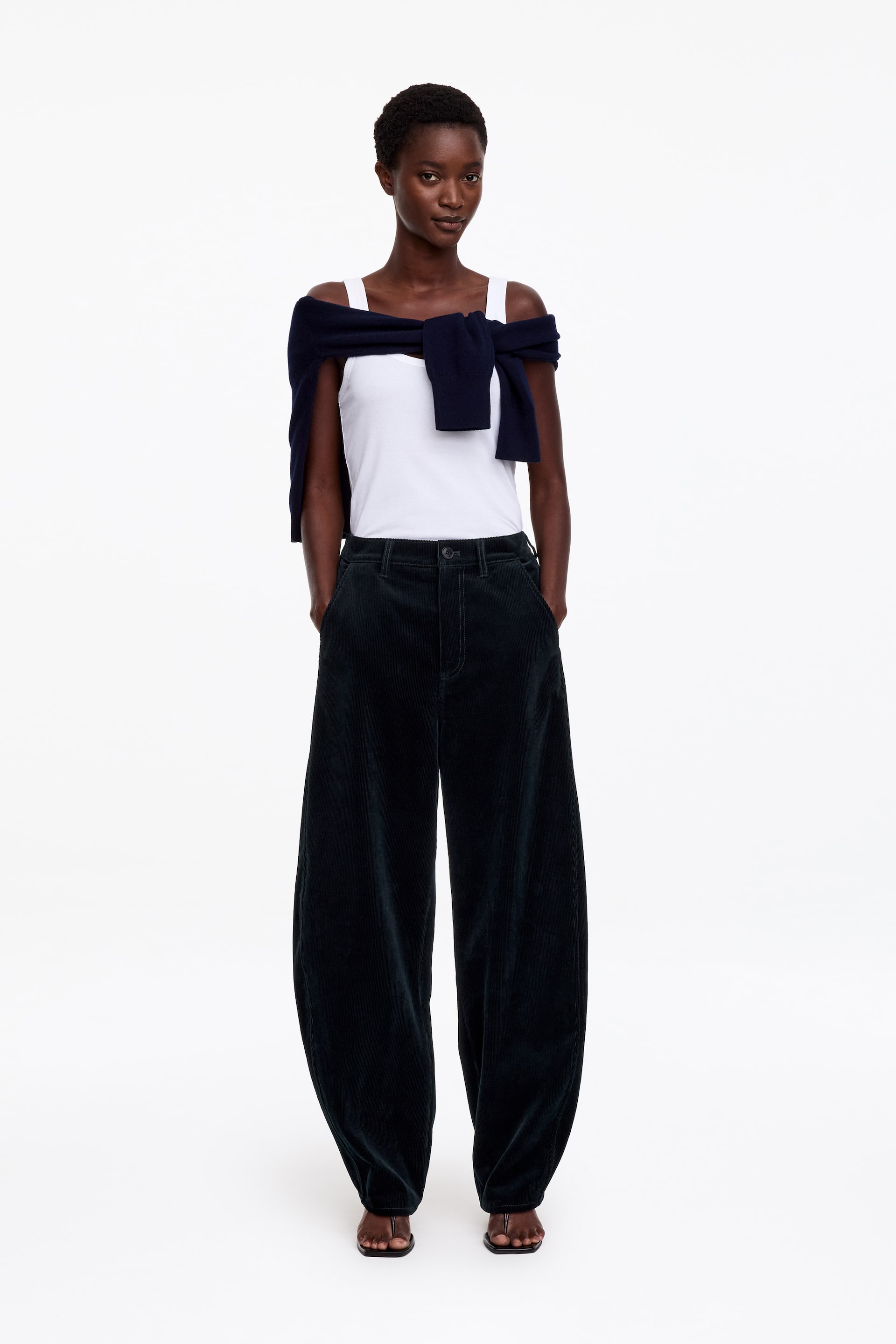 Agrandir l'image: Ce pantalon en velours côtelé foncé, doté d’une silhouette barrel-leg arrondie et d’une taille normale, est porté par le mannequin, debout avec les mains dans les poches avant. Le pantalon est associé à un débardeur blanc et à un pull en maille foncée drapé sur les épaules.