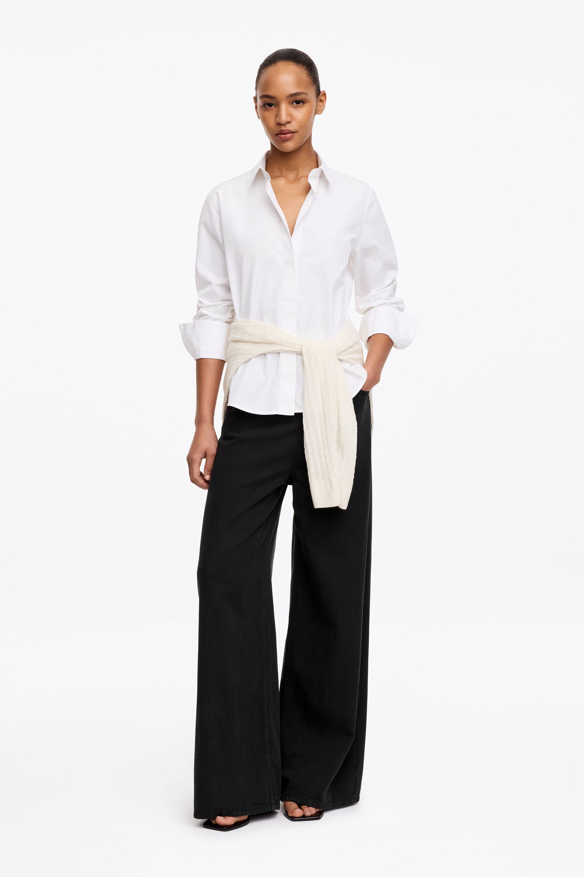 View larger image: The model is wearing black full-length, wide-leg trousers. One hand is placed in a side pocket, and the trousers are styled with a white button-down shirt and a cream sweater tied around the waist.
