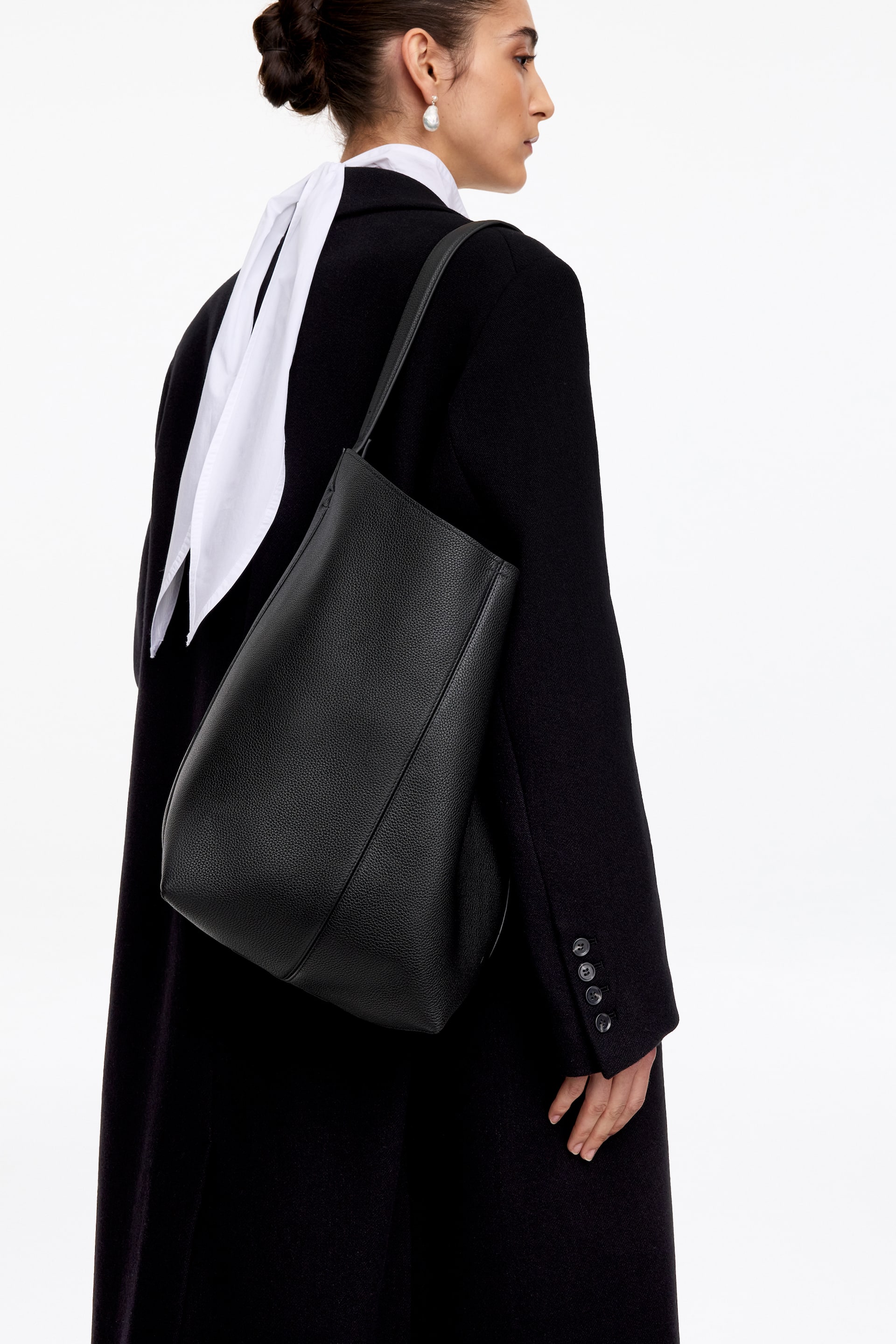 Agrandir l'image: Un cabas en cuir noir grainé à la silhouette souplement structurée est porté à l’épaule par le mannequin. Le sac présente une ligne supérieure sculptée et une fine bandoulière, porté sur un long manteau noir avec une écharpe blanche autour du cou.