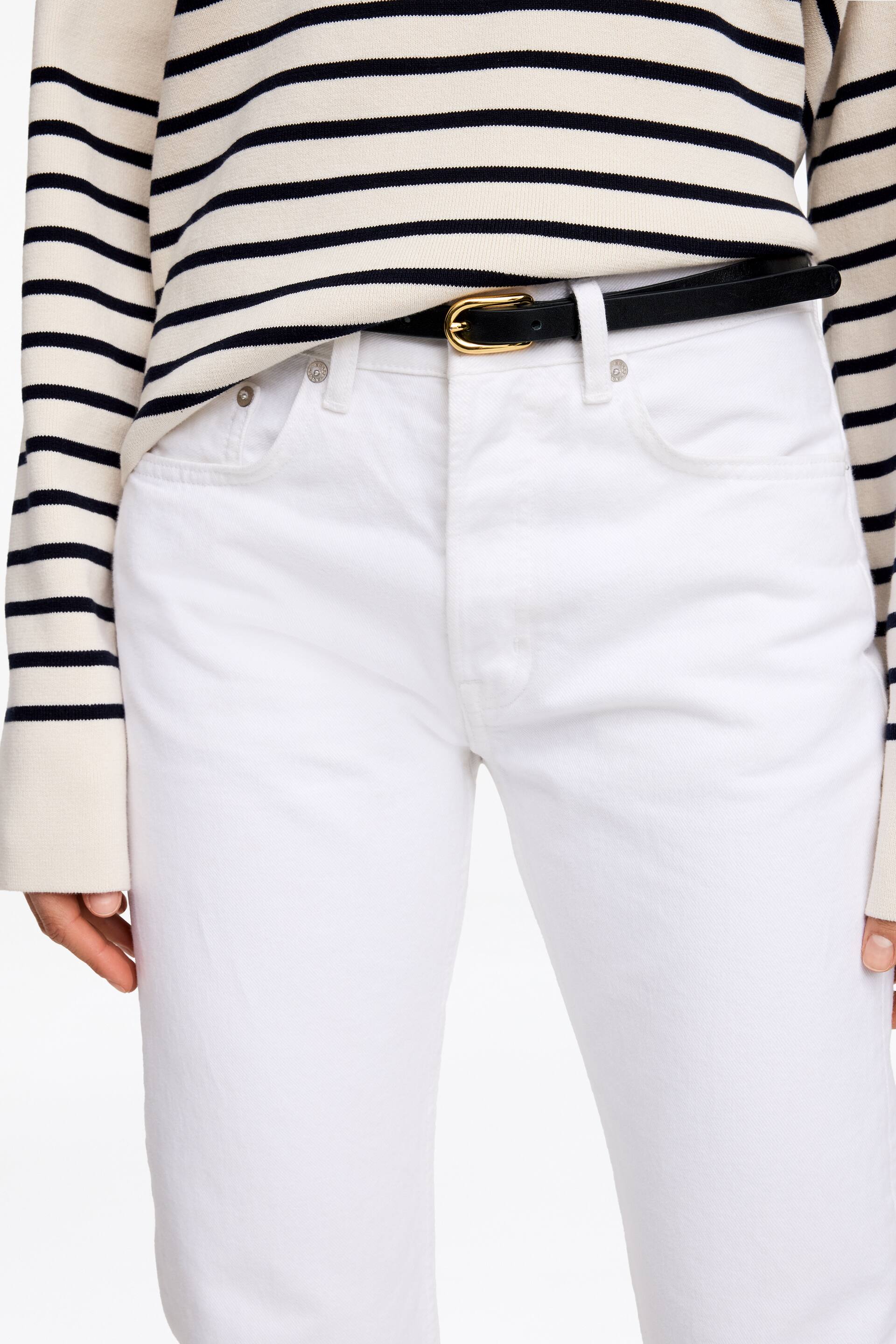 Zobacz większy obraz: Prezentowane są białe jeansy, które mają przednie kieszenie z nitami oraz szlufki na pasek. Czarny pasek z klamrą w złotym odcieniu jest przewleczony przez szlufki, zabezpieczając talię.