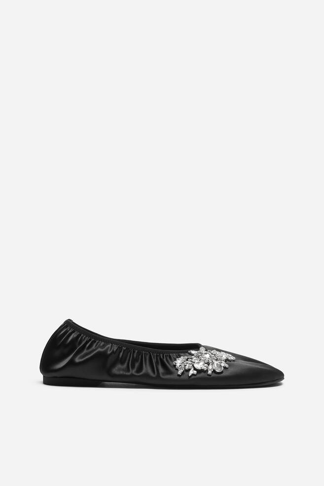 A black satin ballet flat is shown in side profile, featuring a gathered, elasticated collar and a slightly pointed toe. A cluster of clear, sparkling crystals embellishes the side of the toe box. - 1