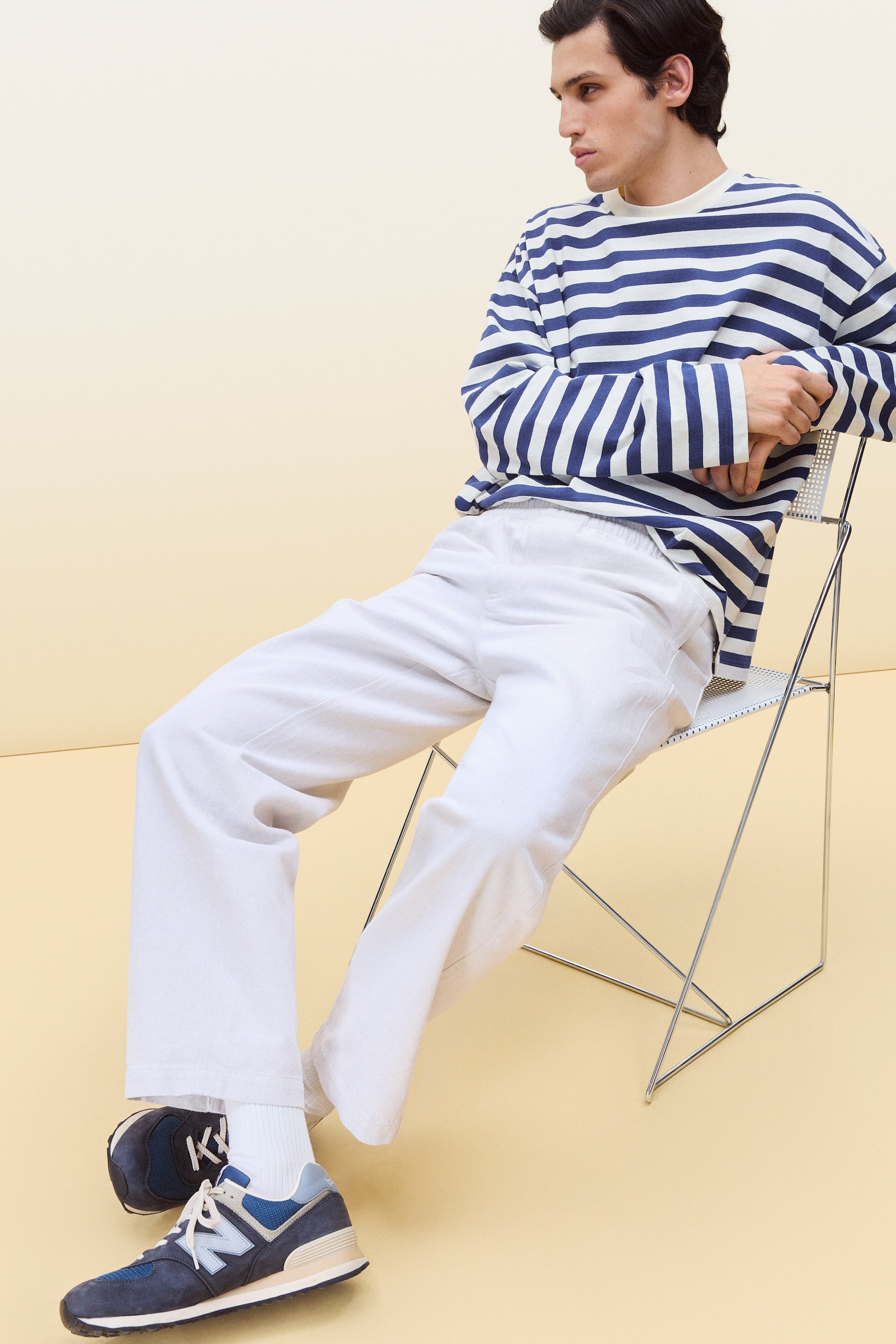 Vis større bilde: En avslappet, oversized langermet topp med hvite og marineblå horisontale striper bæres, med en rund hals. Den er kombinert med lyse beige, vide bukser som faller løst og viser en elastisk midje.