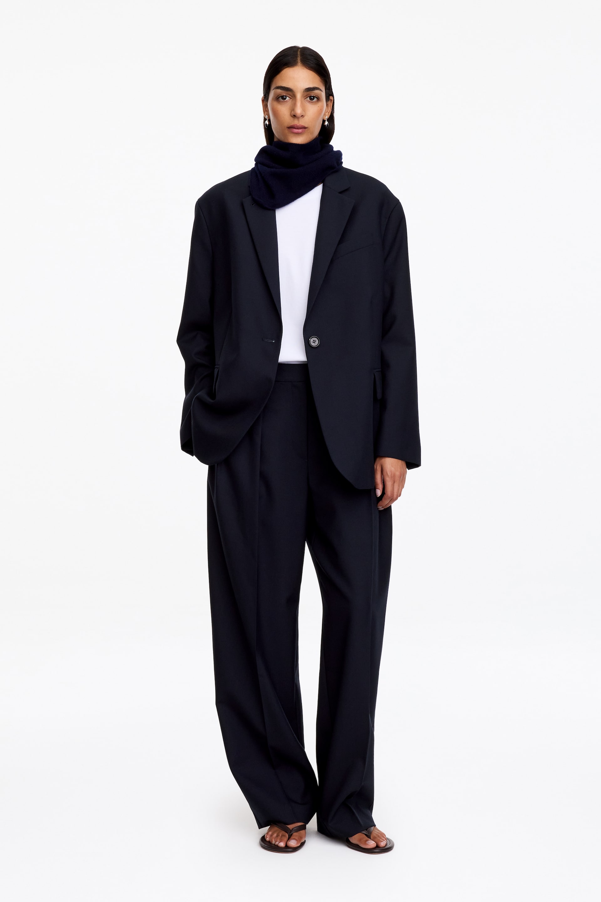 Ver imagem maior: Um blazer azul-marinho de abotoamento simples, de corte descontraído, é usado pela modelo, com ombros marcados, lapelas com entalhe e bolsos com pala. Está sobreposto a um top branco e a uma gola em malha escura, e combinado com umas calças largas a condizer.