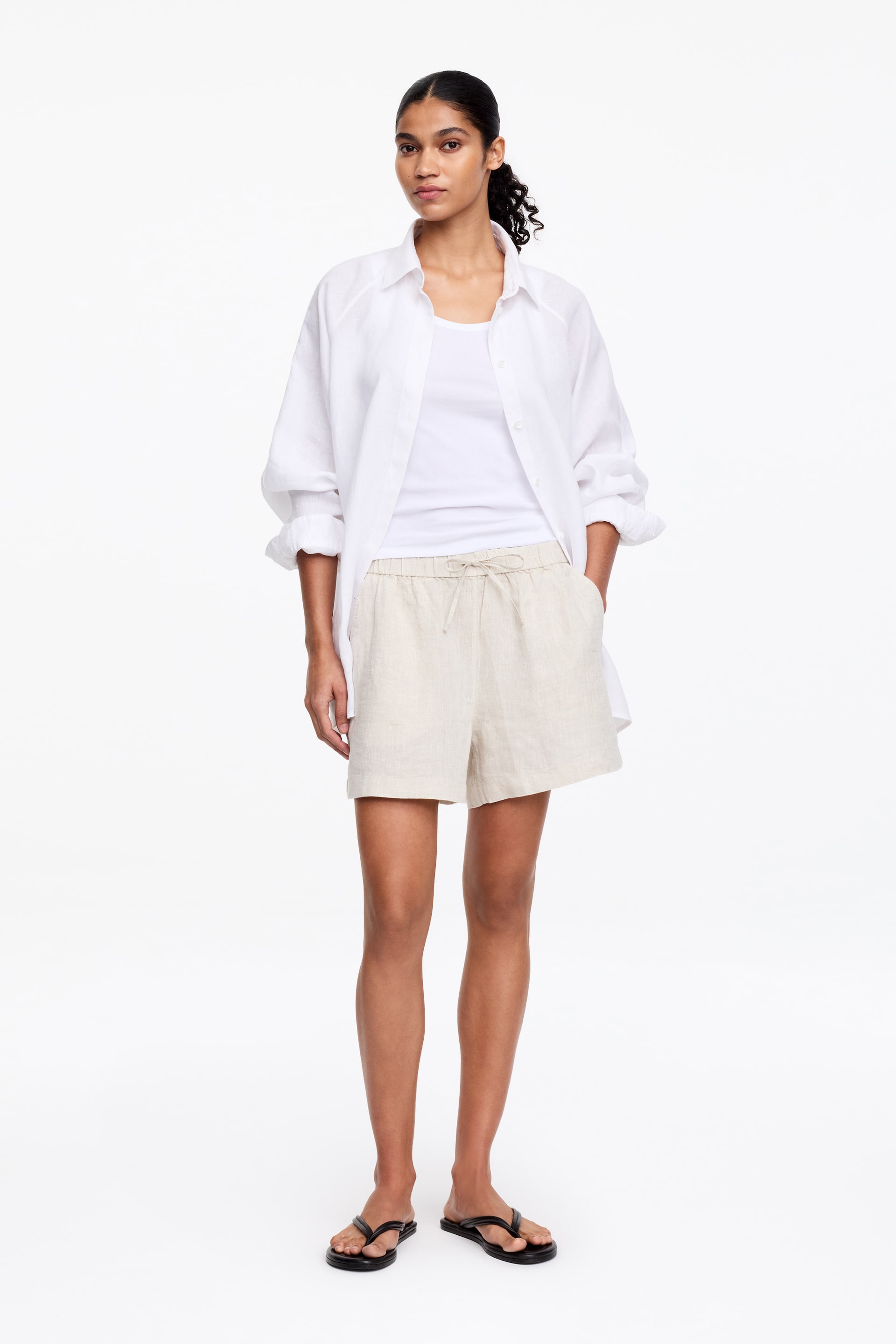 Agrandir l'image: Les shorts beige clair présentent une texture visible semblable au lin, une taille élastiquée avec un cordon de serrage noué à l'avant, et des poches latérales où reposent les mains, offrant une coupe décontractée et se terminant au-dessus du genou.