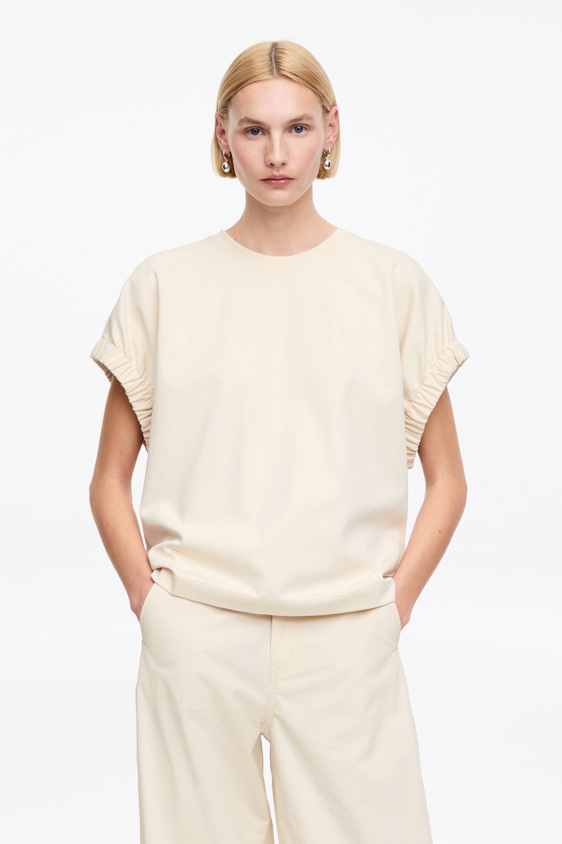Se større billede: Den cremefarvede top, vendt fremad, har en rund halsudskæring, korte ærmer med rynkedetaljer, en afslappet pasform og en lige kant.