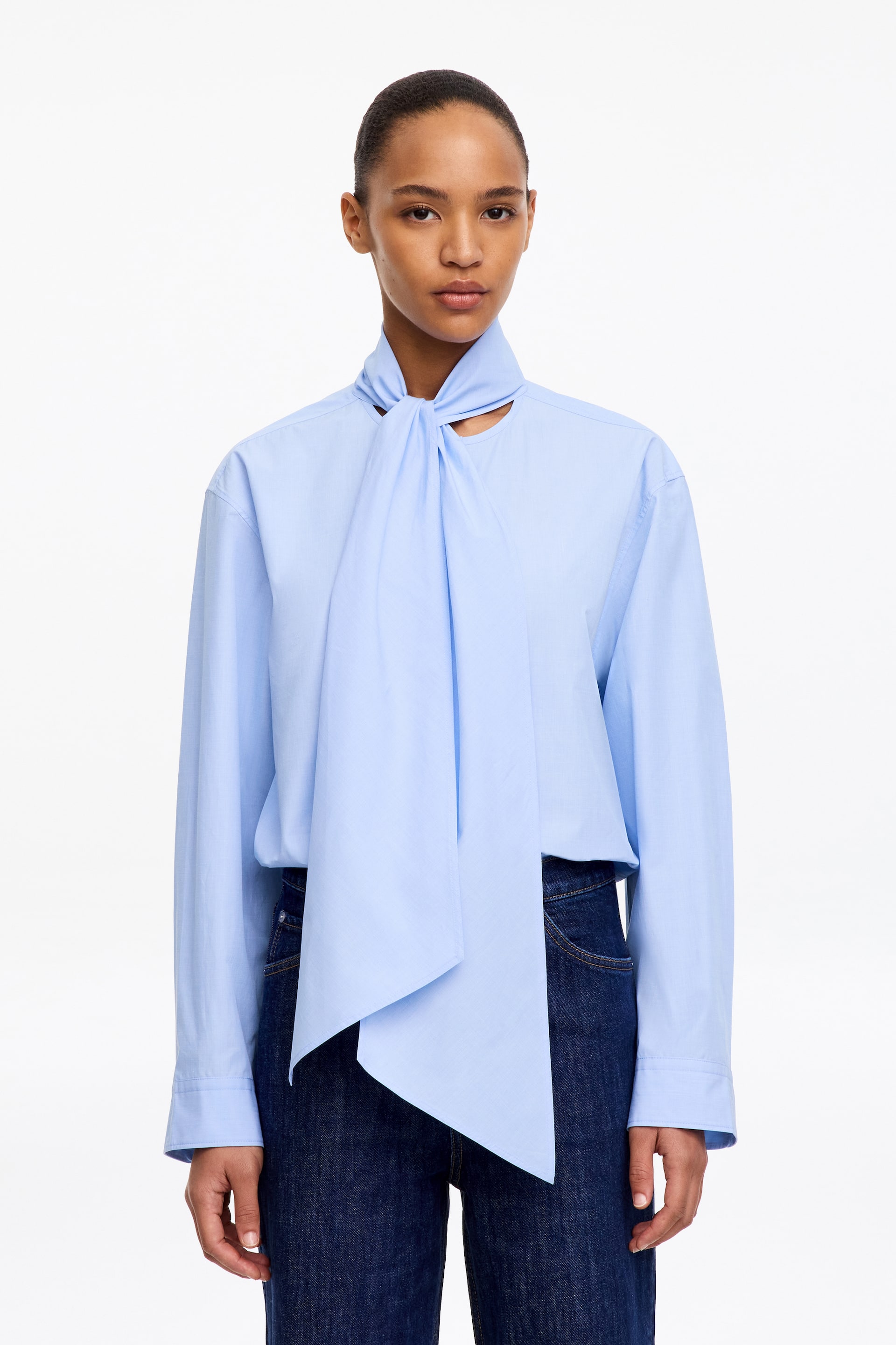 Vezi imaginea mai mare: Bluza cu eșarfă din bumbac Pima, în albastru deschis, este purtată de model. Bluza este stilizată cu o eșarfă integrată, legată la gât, ale cărei capete lungi cad în față, peste blugi de culoare închisă.