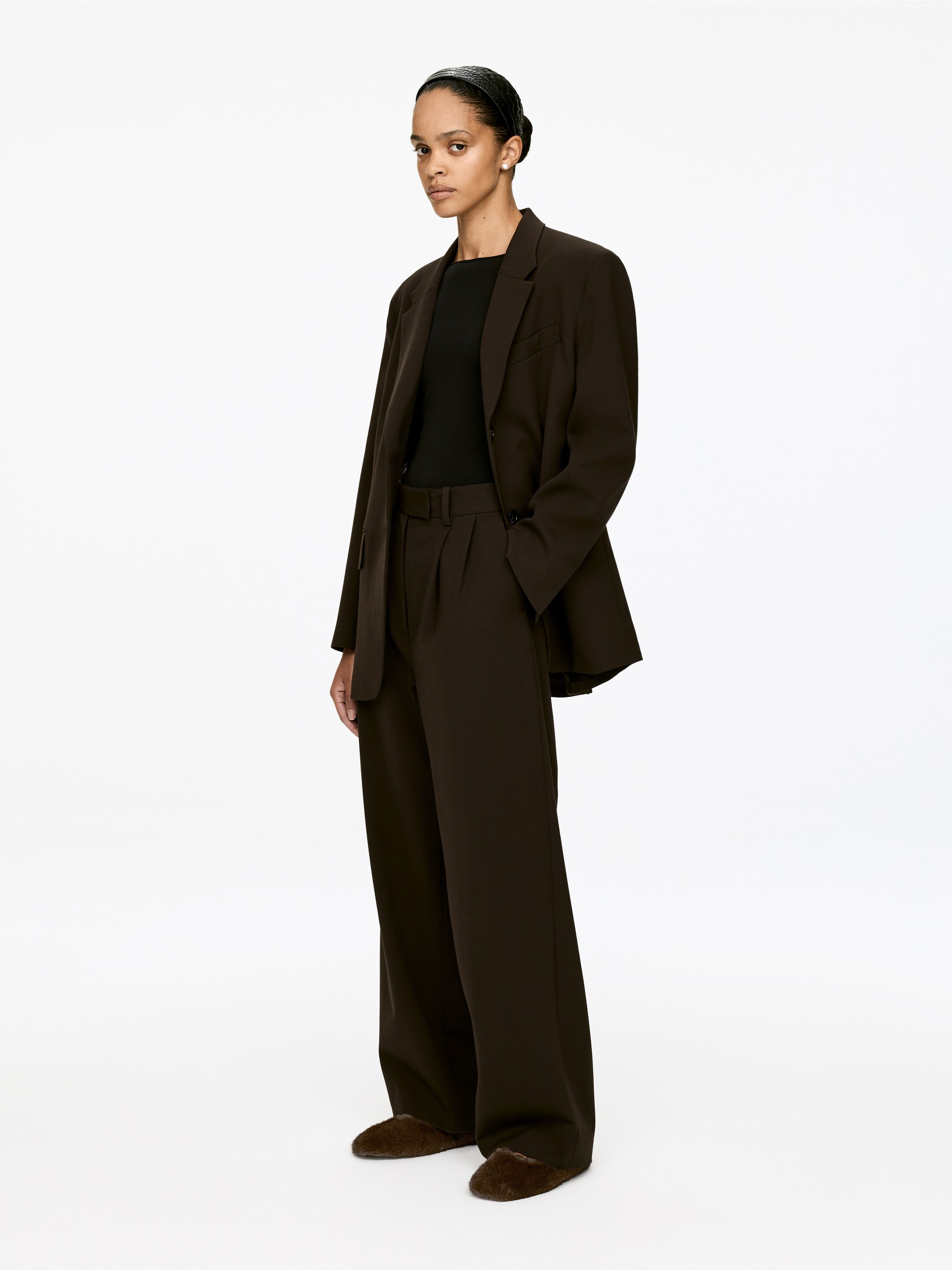 View larger image: A dark brown suit, comprising a relaxed-fit blazer with notch lapels and wide-leg pleated trousers, is being shown from a slight angle to the viewer's left. A black boat-neck top is being worn layered beneath the blazer, and dark brown furry slippers are visible on the feet.