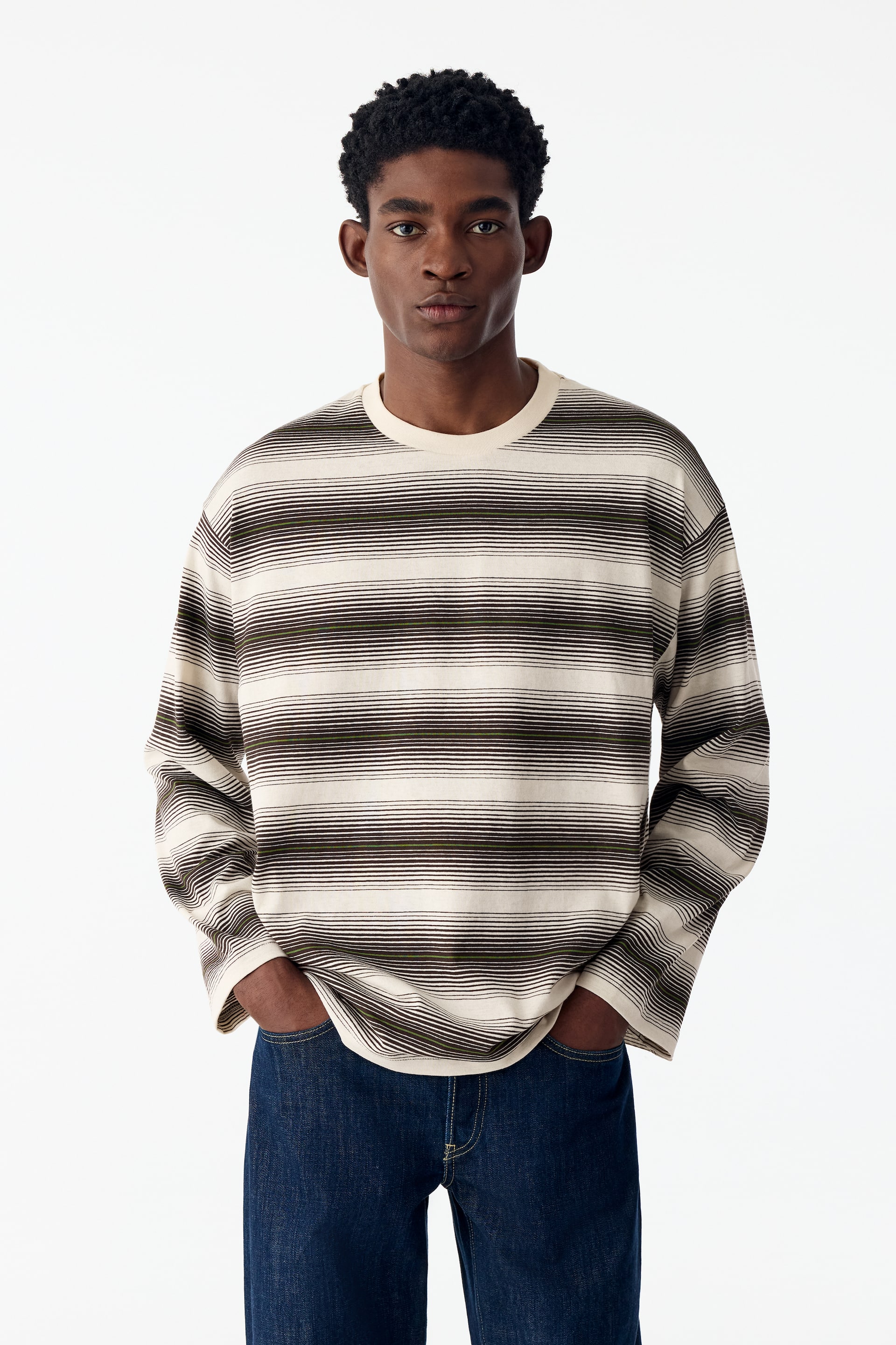 Se større billede: En langærmet T-shirt med rund hals og vandrette striber i creme og mørke farver bæres af modellen. Det afslappede plagg er stylet med mørkeblå jeans, og modellens hænder hviler i lommerne.