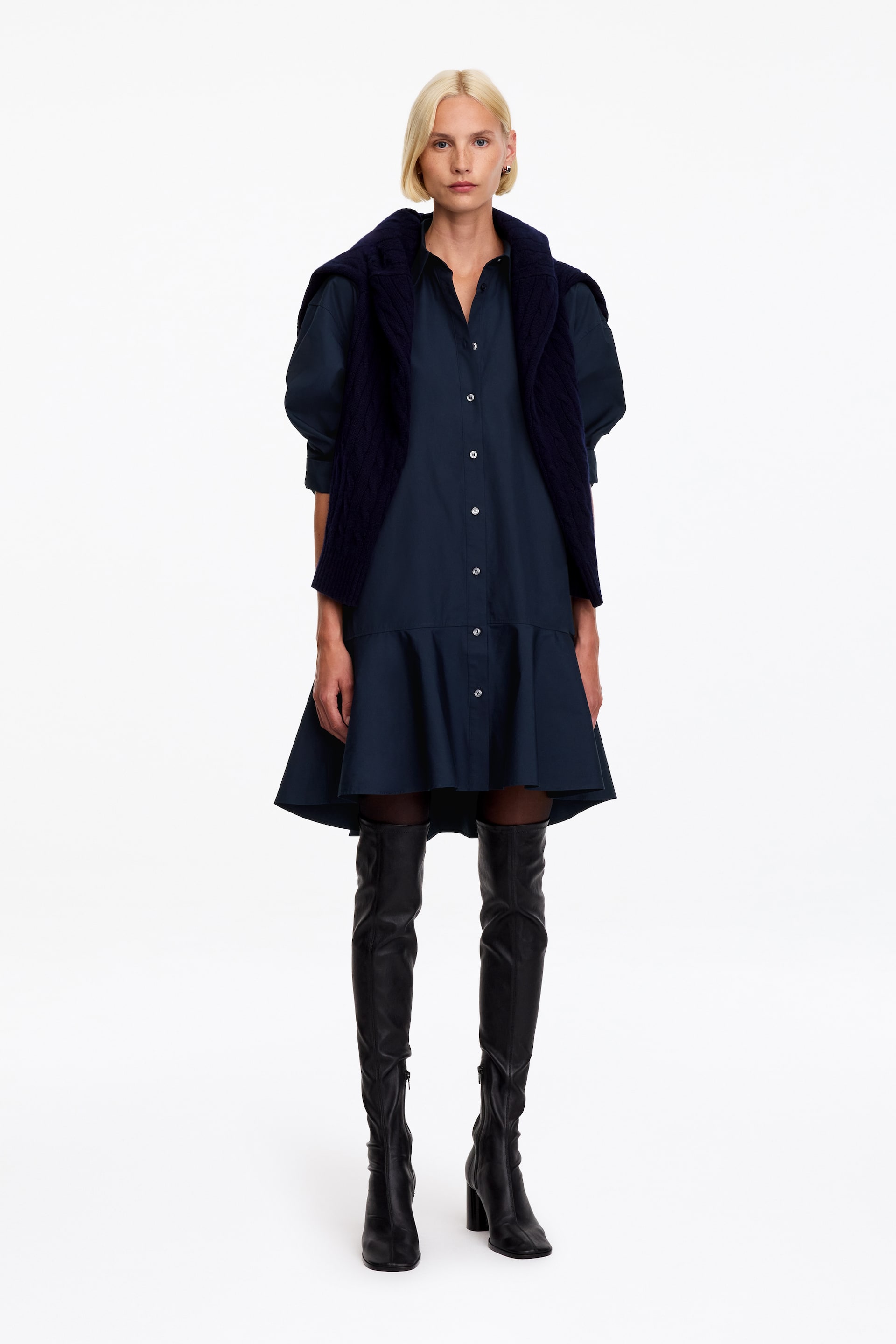 View larger image: The model is wearing a navy blue shirt dress, featuring a classic collar, a button-through front, and a flared, gathered peplum hem. The garment is layered with a dark navy knit sweater draped over the shoulders and styled with black over-the-knee leather boots.