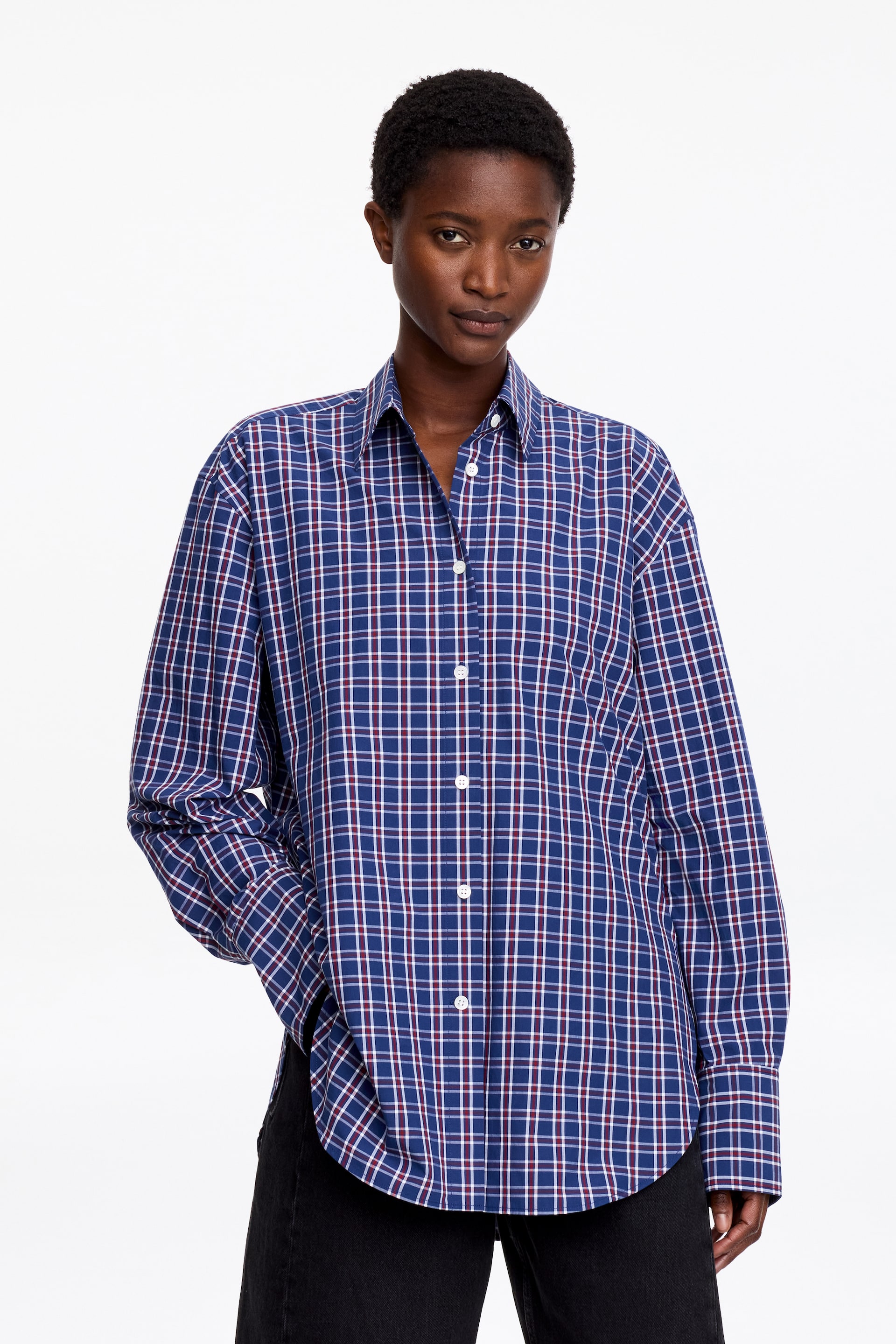 View larger image: A blue, red, and white check button-front shirt is being showcased, featuring a pointed collar, white buttons, and long sleeves with buttoned cuffs. The relaxed-fit shirt displays a curved hem and is styled with black trousers.
