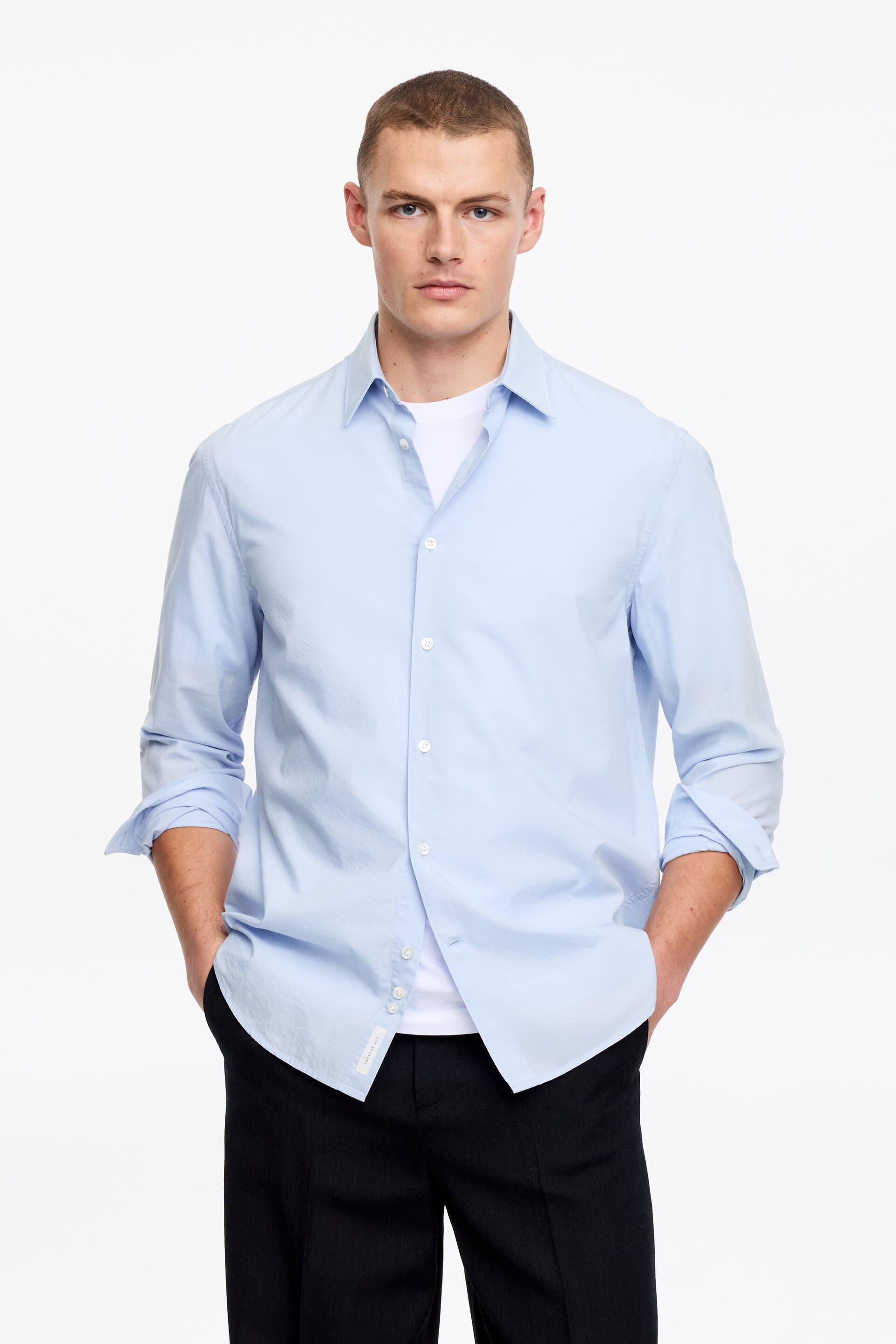 Grotere afbeelding bekijken: Een lichtblauw overhemd met knopen wordt gedragen, naar voren gericht, met een puntige kraag en witte knopen aan de voorkant. De lange mouwen zijn opgerold en het is gelaagd over een wit T-shirt.