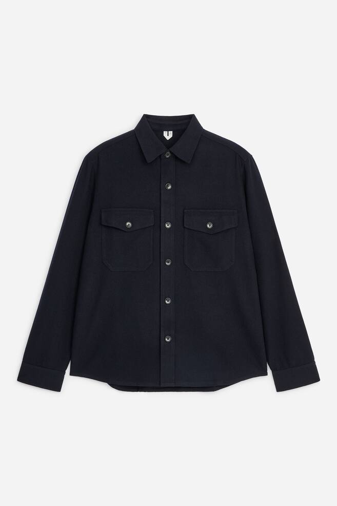 A dark navy blue overshirt is facing the front, featuring a pointed collar, a central button fastening, and two chest patch pockets with buttoned flaps. The long sleeves extend downwards, ending in structured cuffs. - 1