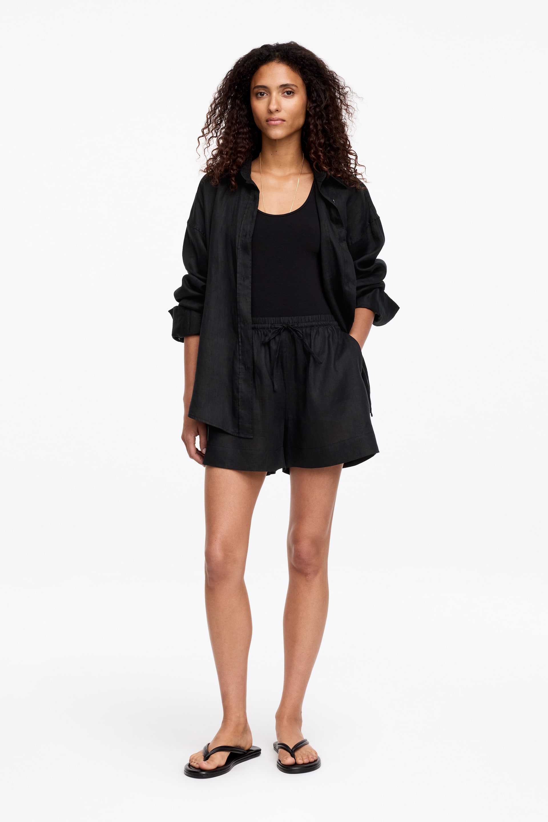 View larger image: A black linen button-up shirt is worn open over a black vest, paired with black linen shorts featuring a drawstring waist. The model stands with hands in the shorts' pockets, wearing black flip-flops.