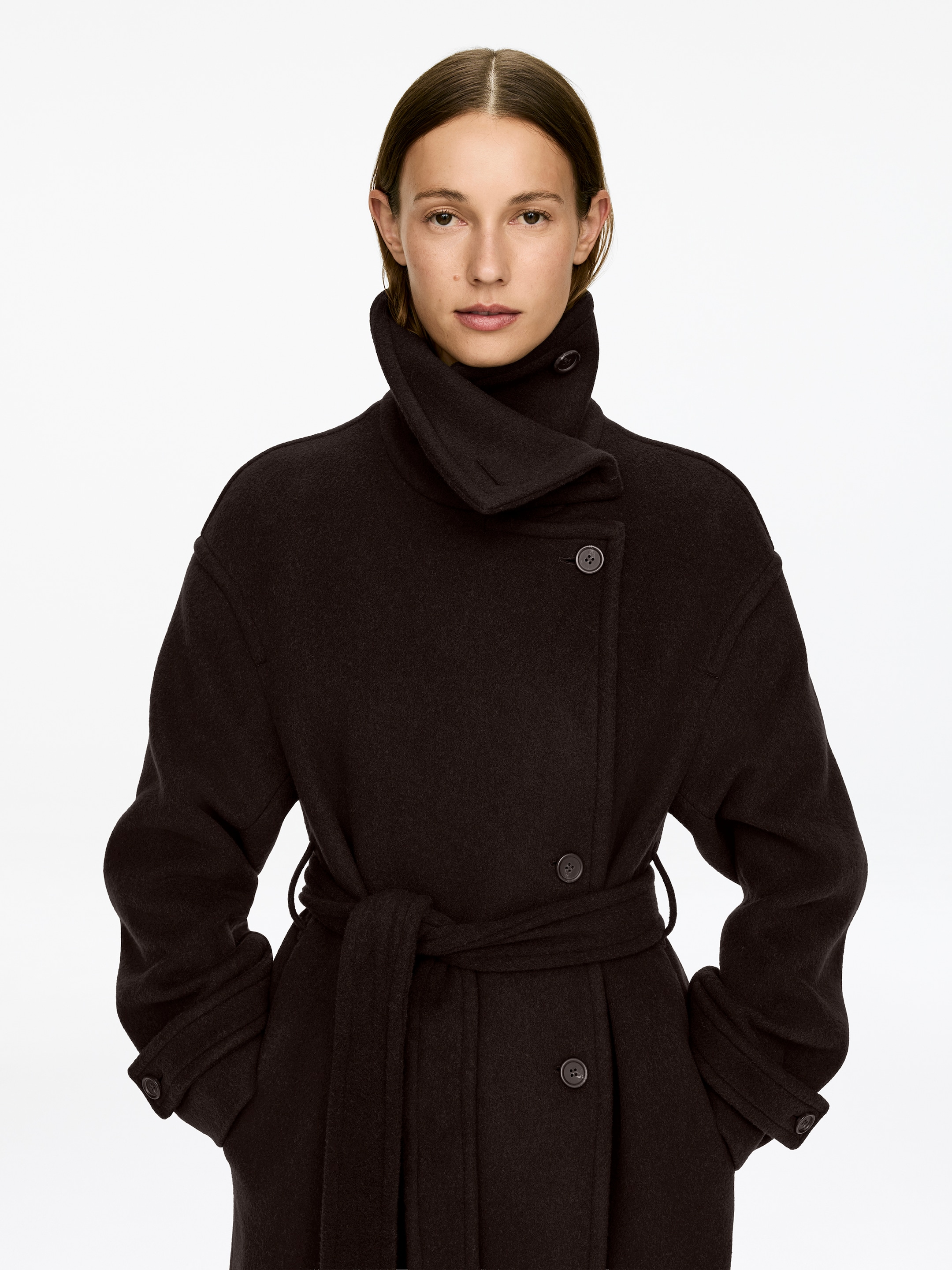 View larger image: An oversized, single-breasted dark brown wool blend belted maxi coat is being worn by the model, featuring a standing collar buttoned at the neck and a belt tied at the waist. The model's hands are placed in the coat's side pockets, and sleeve tabs with button closures are visible at the cuffs.