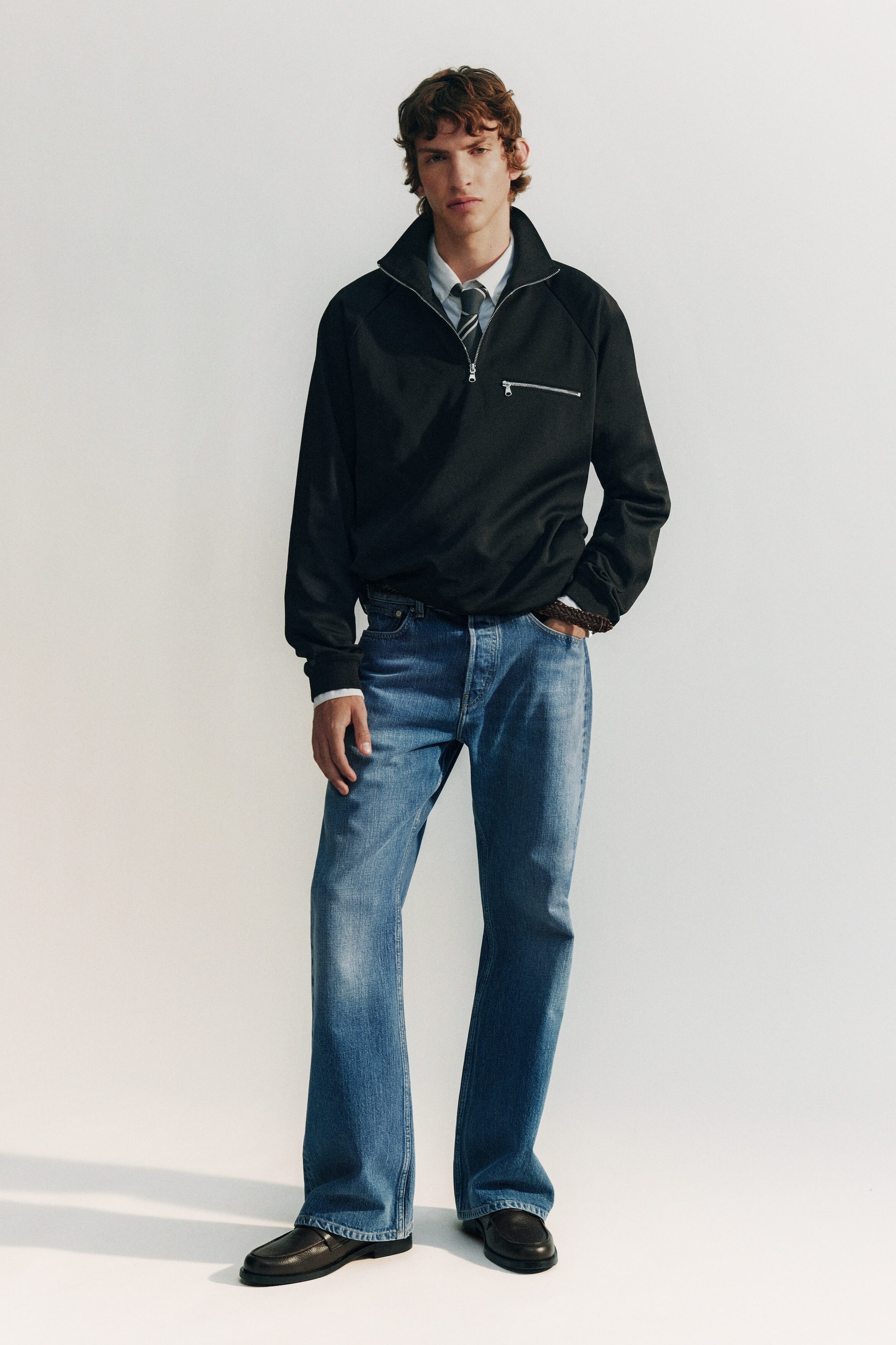 Visualizza immagine più grande: Un pullover nero con zip a un quarto è presentato frontalmente, caratterizzato da un colletto rialzato e una zip color argento che si estende fino al petto superiore. Una tasca orizzontale con zip color argento è visibile sul petto sinistro del portatore, e il pullover è indossato sopra una camicia di colore chiaro e una cravatta a righe.