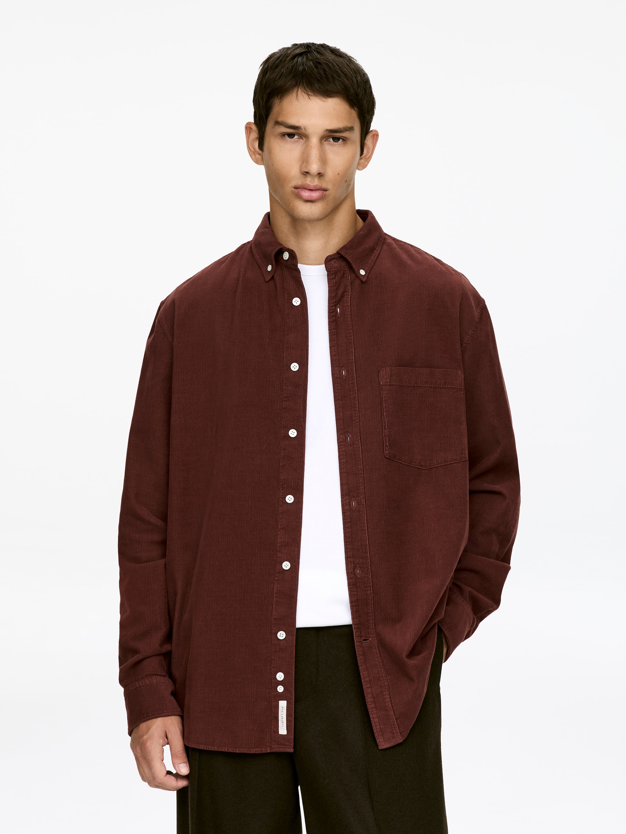 Agrandir l'image: Une chemise en velours côtelé bordeaux orientée vers l'avant est présentée, avec un col boutonné, une patte de boutonnage complète avec des boutons blancs, et une seule poche plaquée sur la poitrine gauche. Elle est portée ouverte sur un t-shirt blanc.