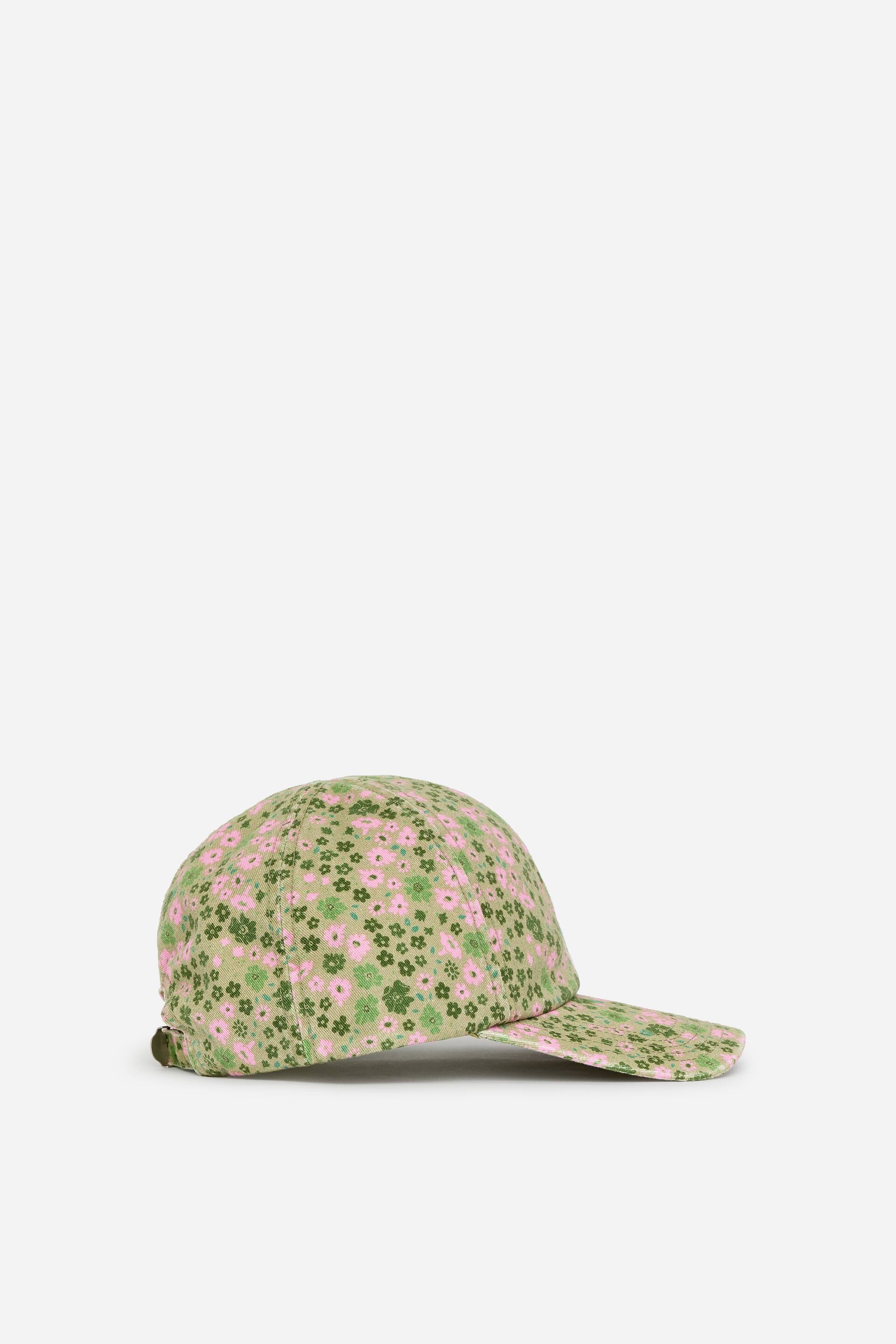 Vis større bilde: En baseballcap er vinklet til siden, og viser en olivengrønn stoffbase mønstret med rosa og mørkegrønne blomster. Den har en lett buet brem og en justerbar stropp med et synlig spenne bak.