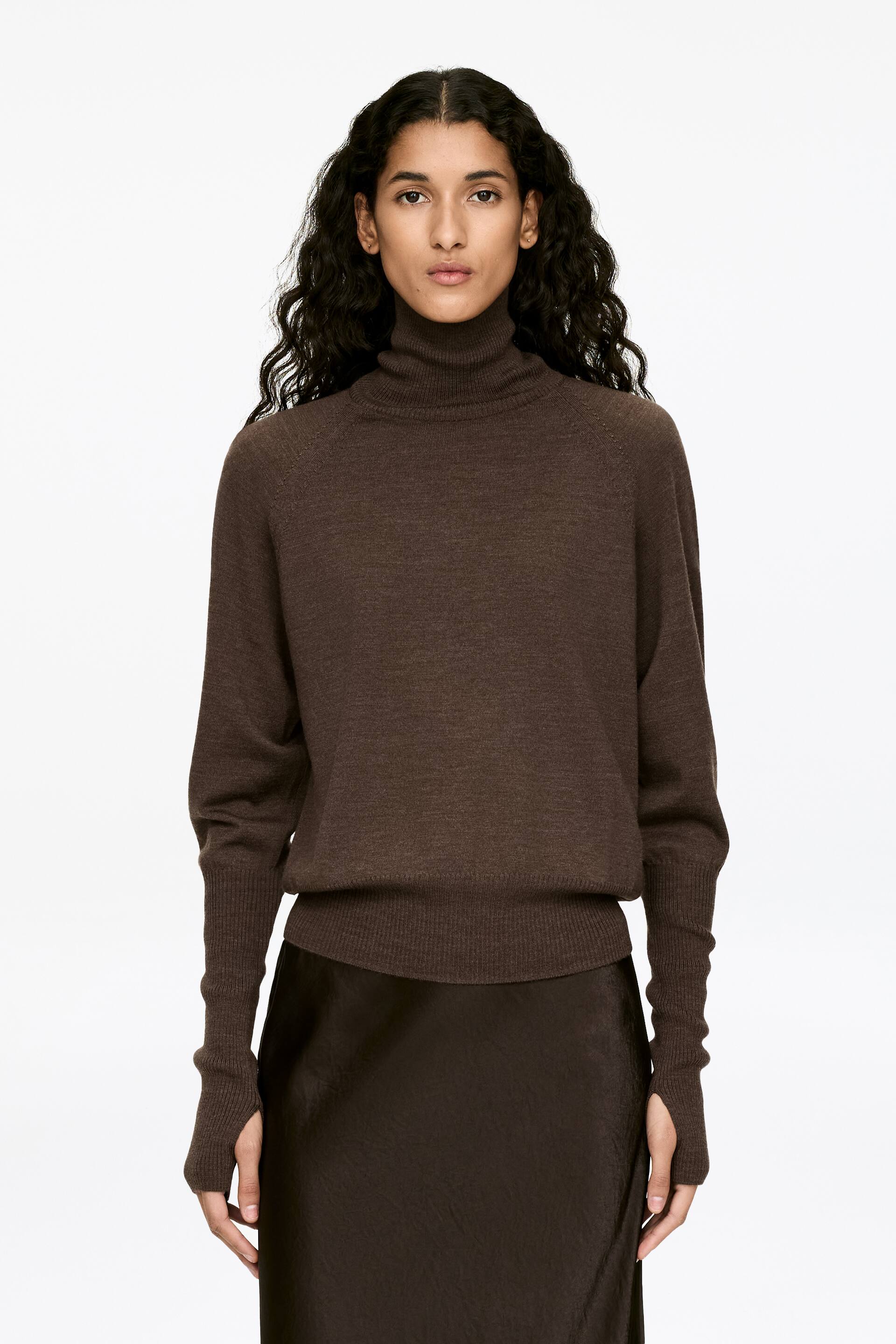Größeres Bild ansehen: Ein dunkelbrauner Rollkragenpullover mit gerippten Abschlüssen am Rollkragen, Saum und verlängerten Bündchen wird vom Modell getragen. Die Hände des Modells ragen durch Daumenlöcher in den Bündchen, und der Pullover wird über einem dunkelbraunen Rock getragen.