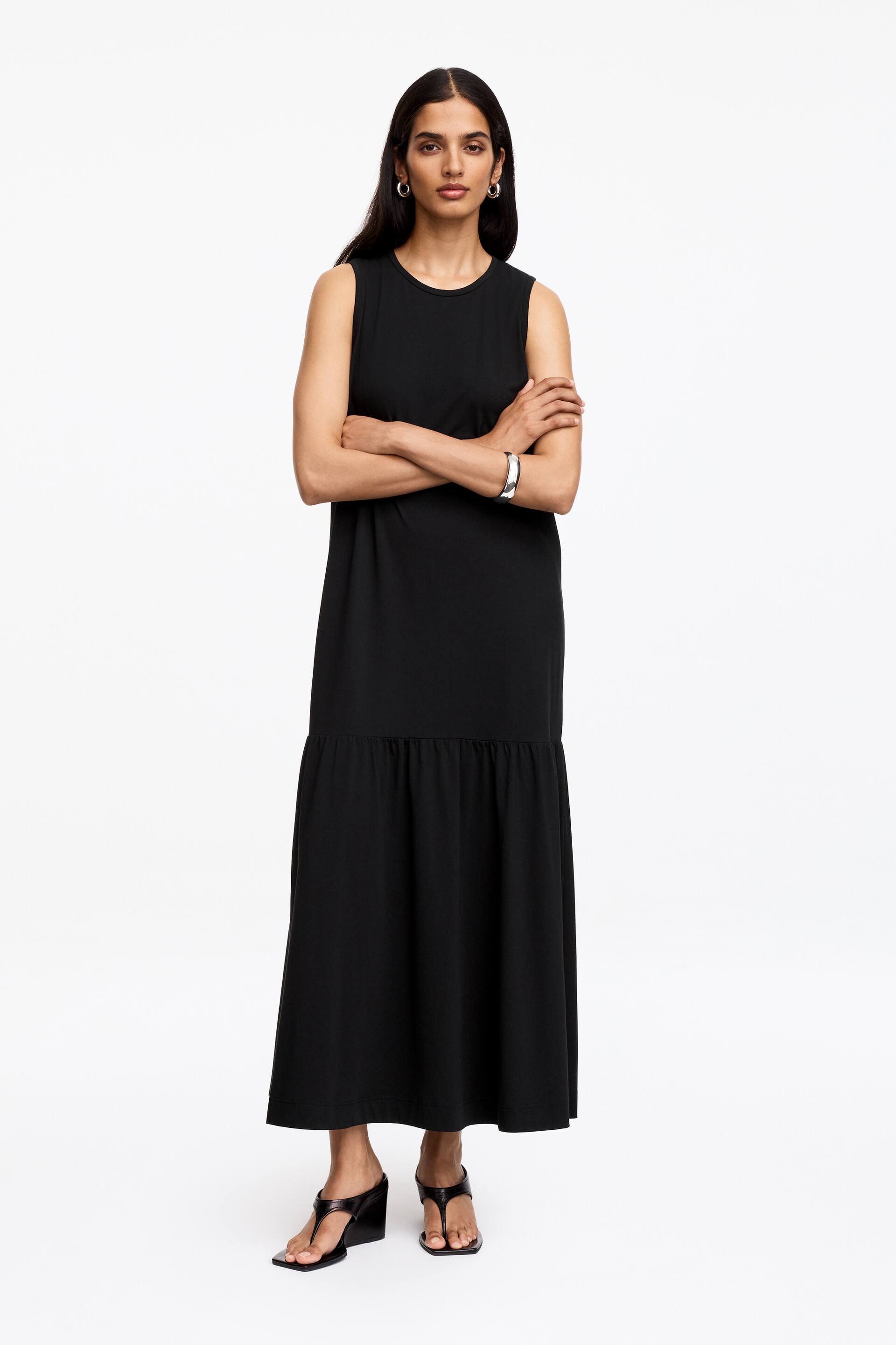 Größeres Bild ansehen: Ein schwarzes ärmelloses Maxikleid wird von vorne gezeigt, mit einem runden Ausschnitt und einer gerafften Naht, die einen stufigen Rock am oberen Oberschenkel bildet und bis zur Knöchellänge reicht.