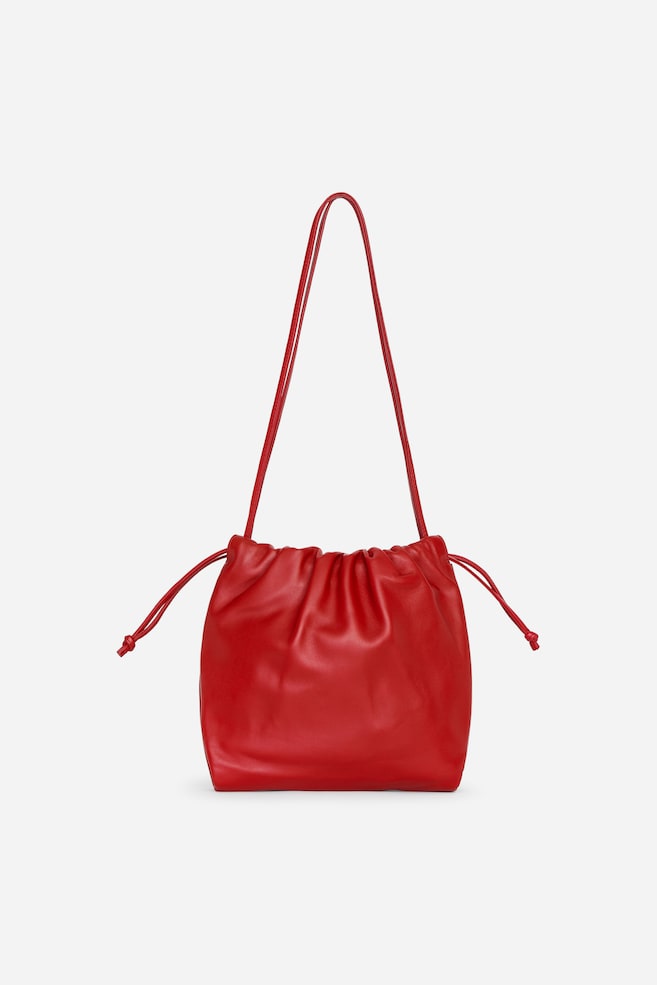 Un sac bandoulière en cuir rouge est présenté de face, avec une silhouette souple façon bourse et une texture douce et décontractée. Le modèle présente de longues bandoulières fines en cuir qui remontent vers le haut, ainsi que des cordons de serrage coordonnés, dont les extrémités nouées dépassent de chaque côté. - 1