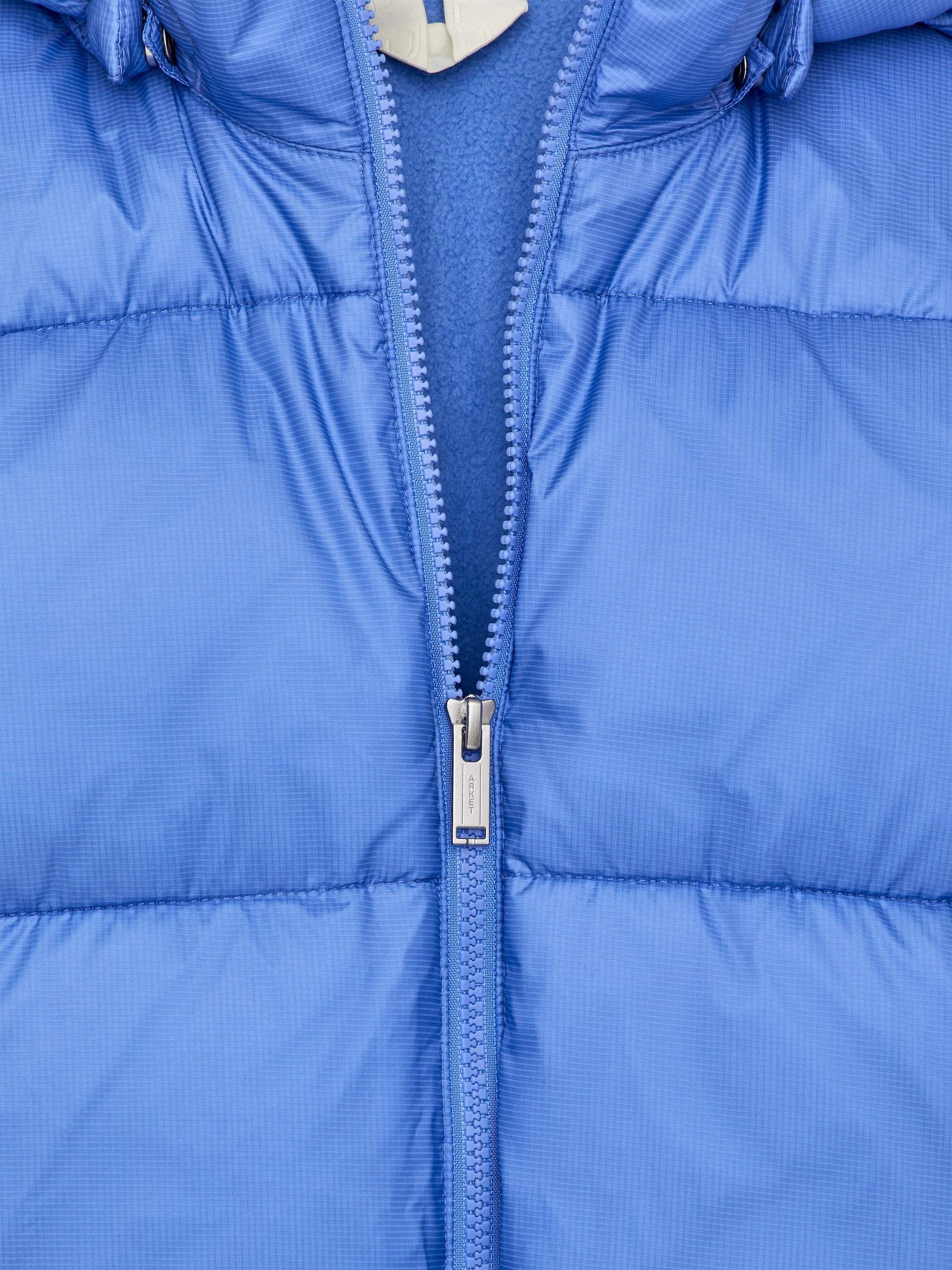Agrandir l'image: Une veste matelassée bleue est présentée de face, avec une fermeture éclair partiellement ouverte. La fermeture éclair révèle une doublure en polaire bleue, tandis que des lignes de matelassage horizontales sont visibles sur toute la veste, et une capuche est attachée en haut.