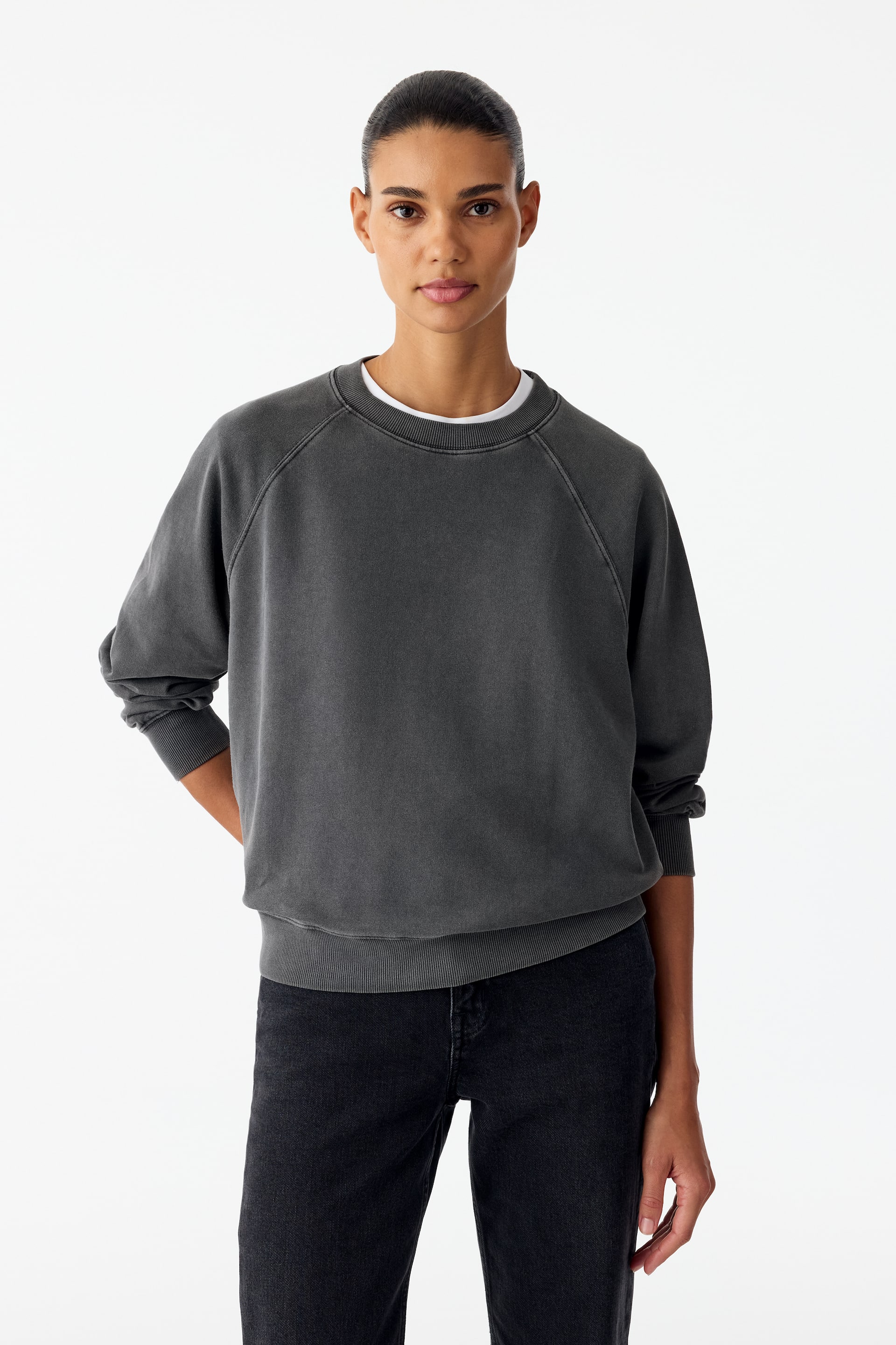 Ver imagem maior: Um sweatshirt de gola redonda em cinzento escuro, com lavagem vintage desbotada, está a ser usado pelo modelo. Apresenta mangas raglan e acabamentos canelados na gola, nos punhos e na bainha, e está sobreposto a uma gola de t-shirt branca, conjugado com jeans pretos.