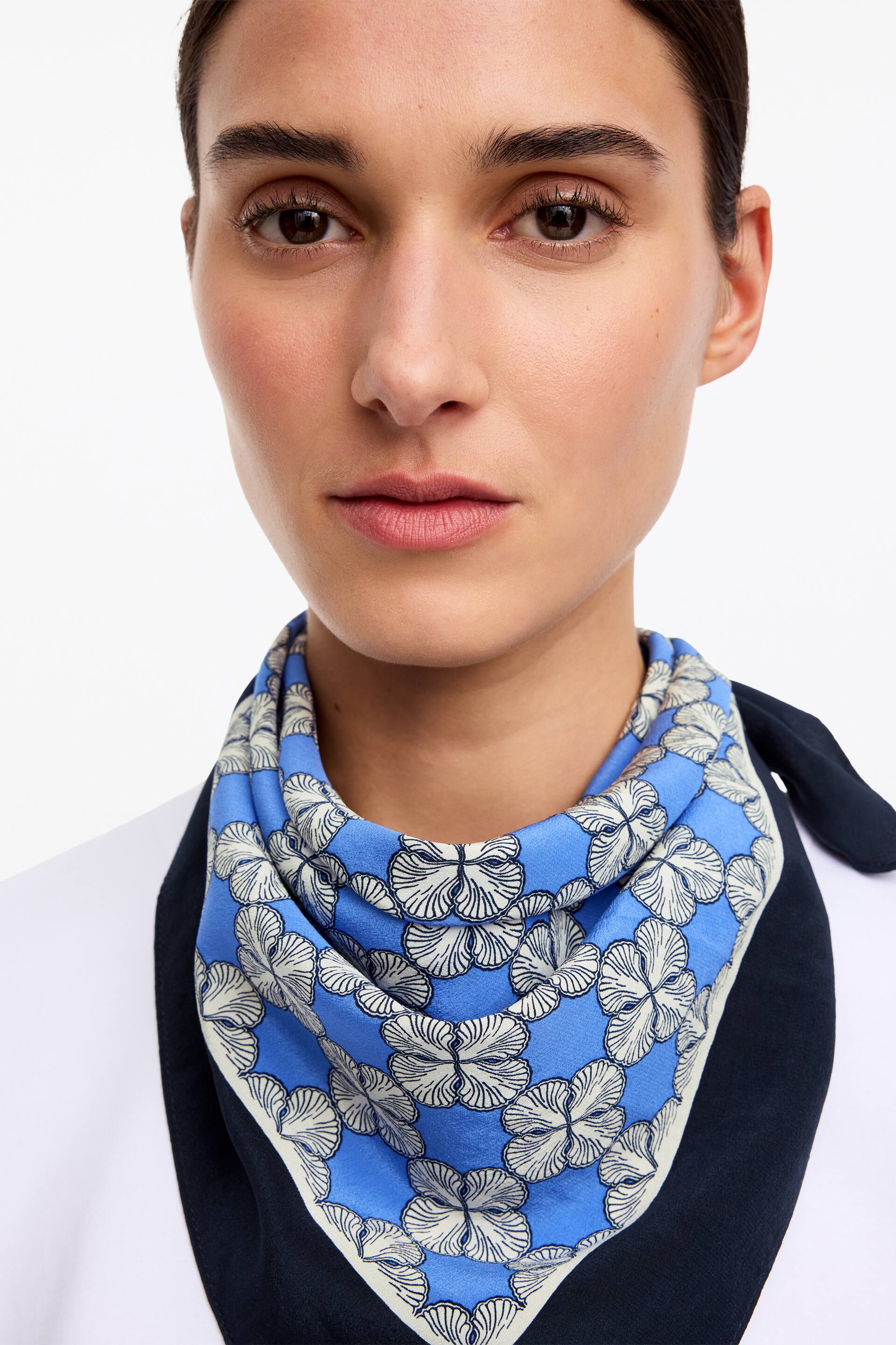Nagyobb kép megtekintése: Egy 62 x 62 cm-es selyemsál van a modell nyakán. Kék alapú, fehér és sötétkék virágmintával díszített, nyakkendőként van viselve egy fehér felső és egy sötét ruhadarab fölött.