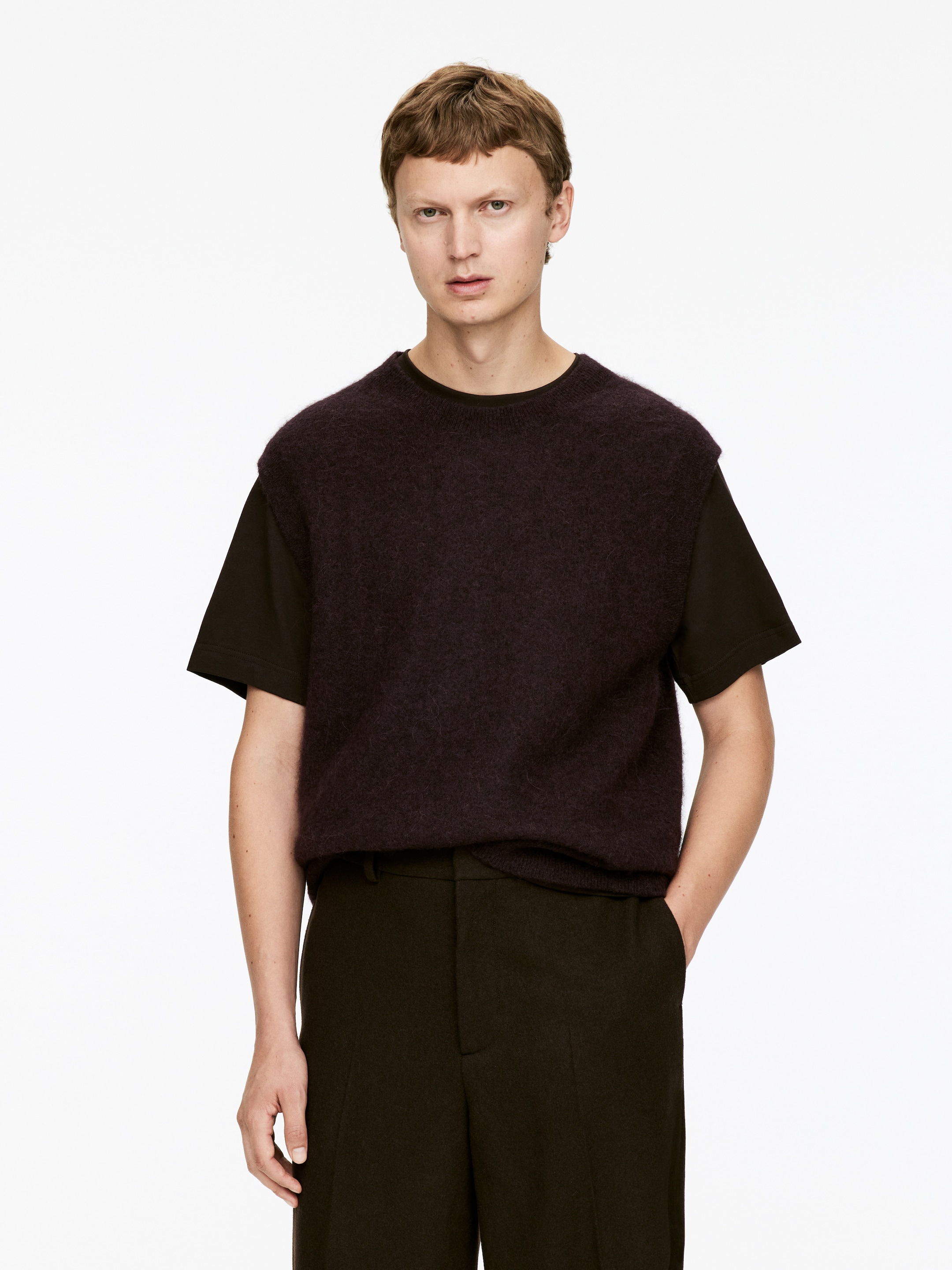 Grotere afbeelding bekijken: Een donker auberginekleurige, pluizige, mouwloze trui wordt gedragen, gelaagd over een donkerbruin t-shirt met korte mouwen. De trui en het t-shirt worden gecombineerd met donker olijfgroene broek, alles gepresenteerd op een frontaal figuur.