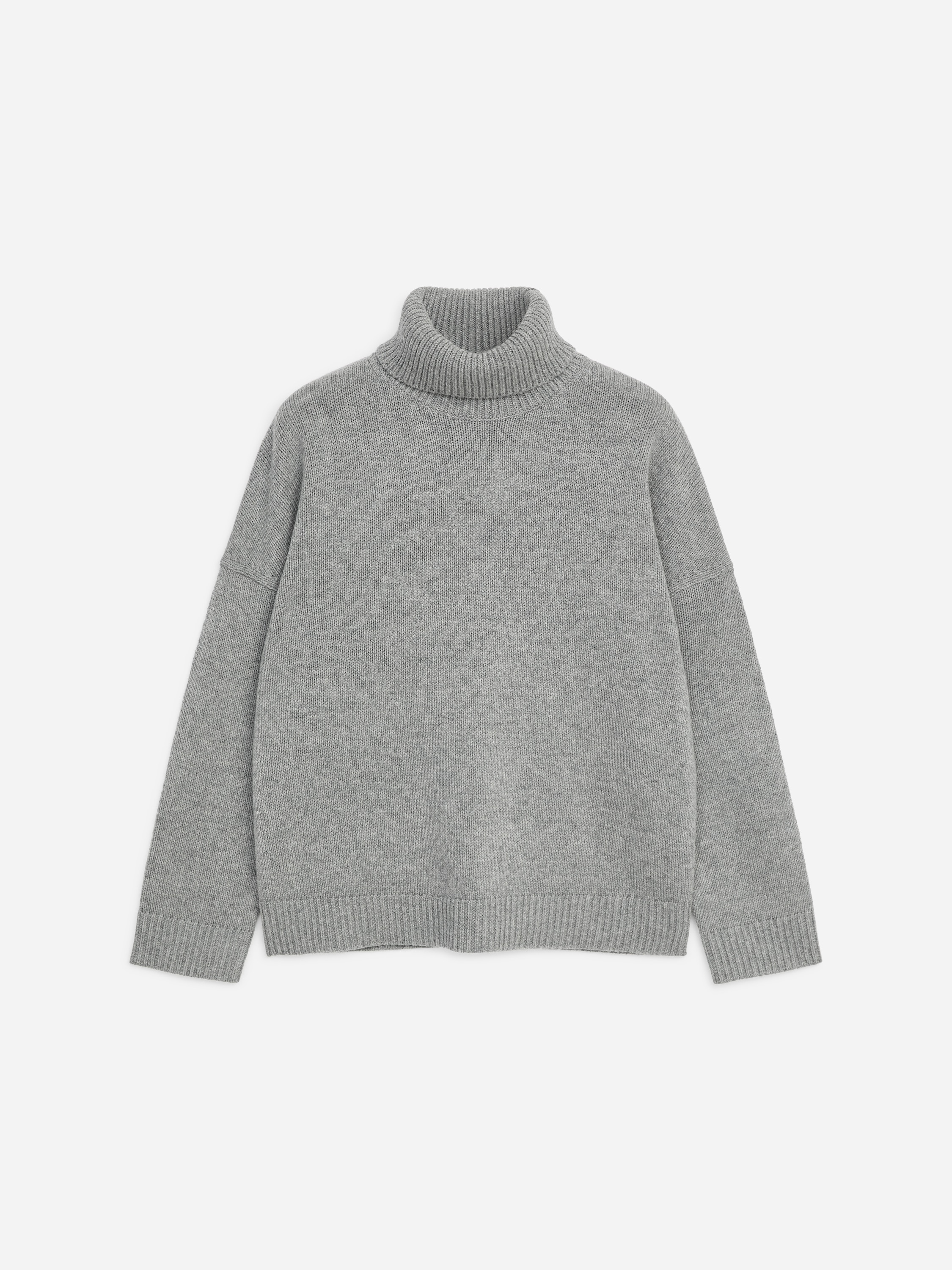 A grey roll-neck wool jumper featuring a relaxed fit, dropped shoulders, and ribbed trims is being worn by the model. The model is standing with hands placed in the pockets of dark trousers.
