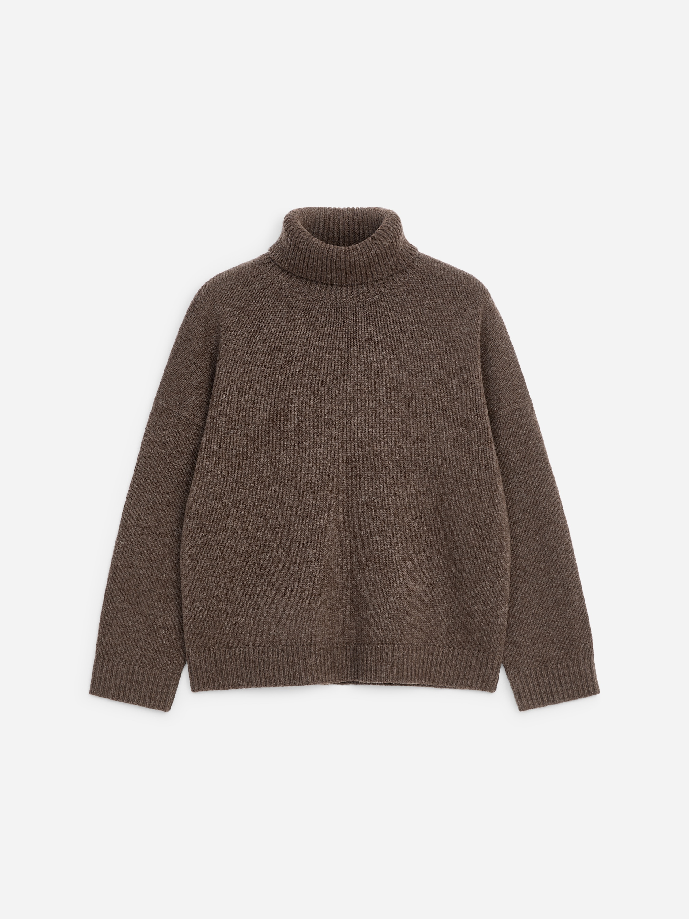 A brown, relaxed-fit roll-neck jumper made of textured wool is being worn by the model, featuring a high ribbed collar, dropped shoulders, and ribbed cuffs and hem. The jumper is paired with off-white trousers.
