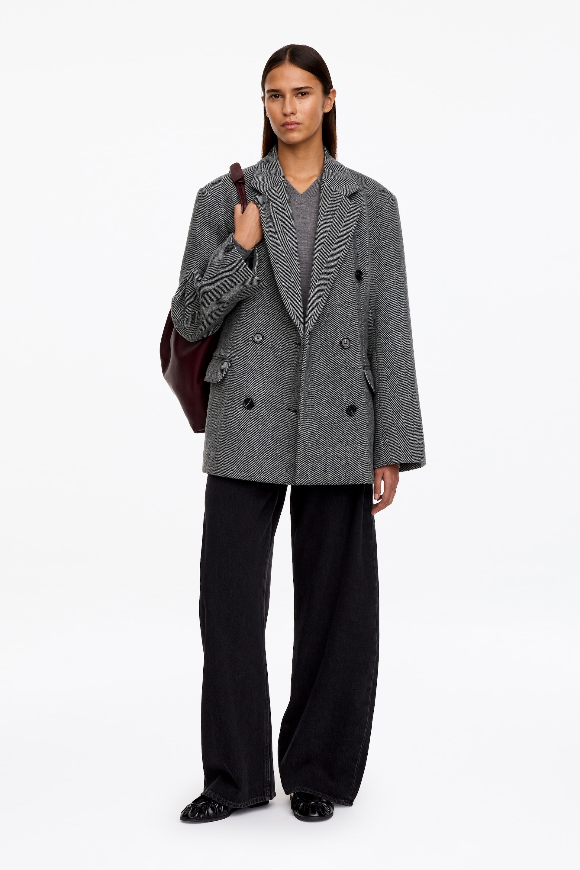 Visualizza immagine più grande: La modella indossa un blazer doppiopetto grigio dalla vestibilità rilassata in tessuto spinato, caratterizzato da revers a lancia, tasche con patta e bottoni scuri. Il blazer è abbinato a un top grigio con scollo a V e a pantaloni neri a gamba larga, mentre la modella tiene la tracolla di una borsa a spalla rosso scuro.