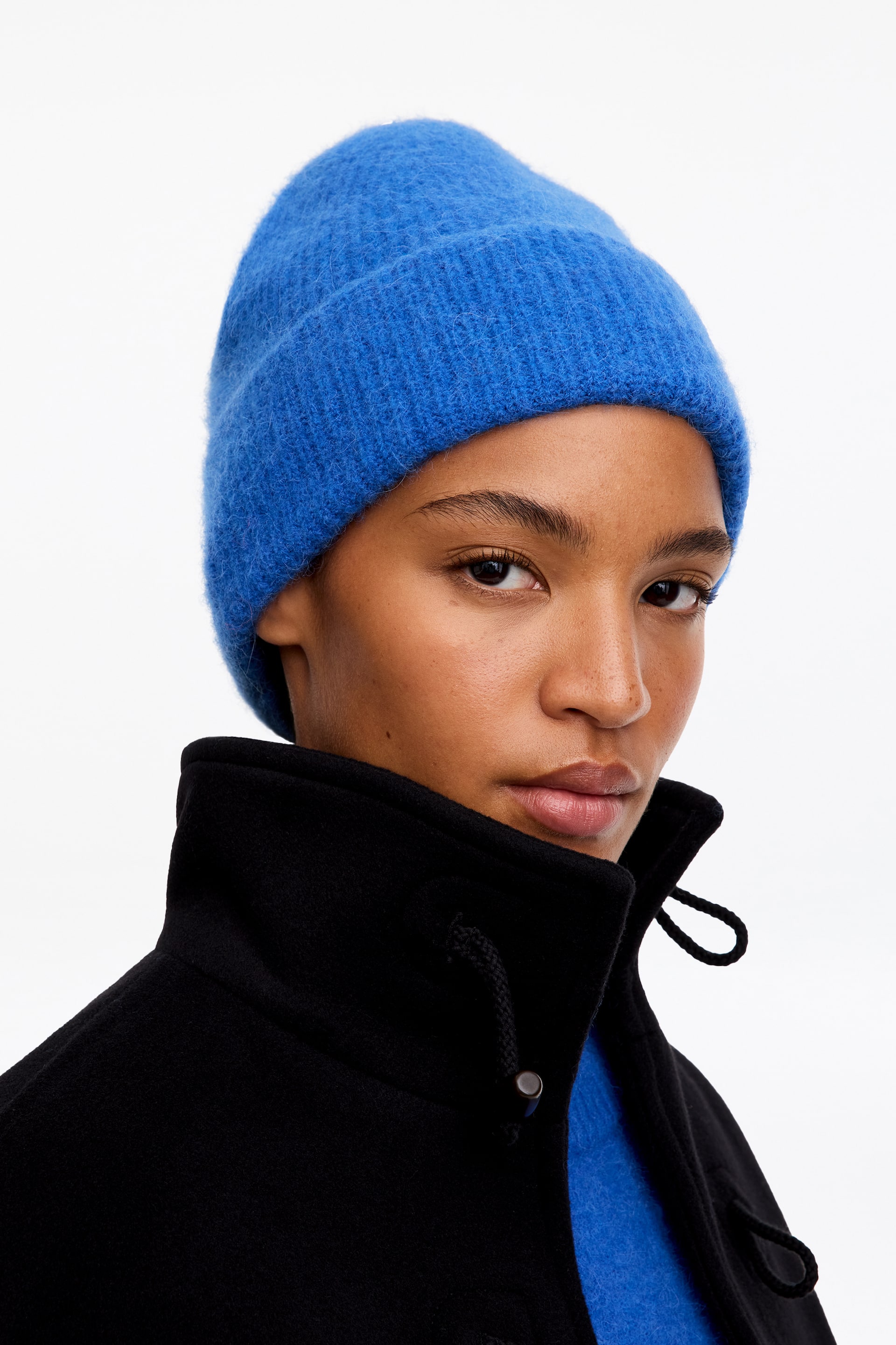 Grotere afbeelding bekijken: Een felblauwe gebreide beanie met een pluizige, geribbelde textuur en omgeslagen boord wordt door het model gedragen. Het accessoire is gestyled met een zwarte jas met hoge kraag.