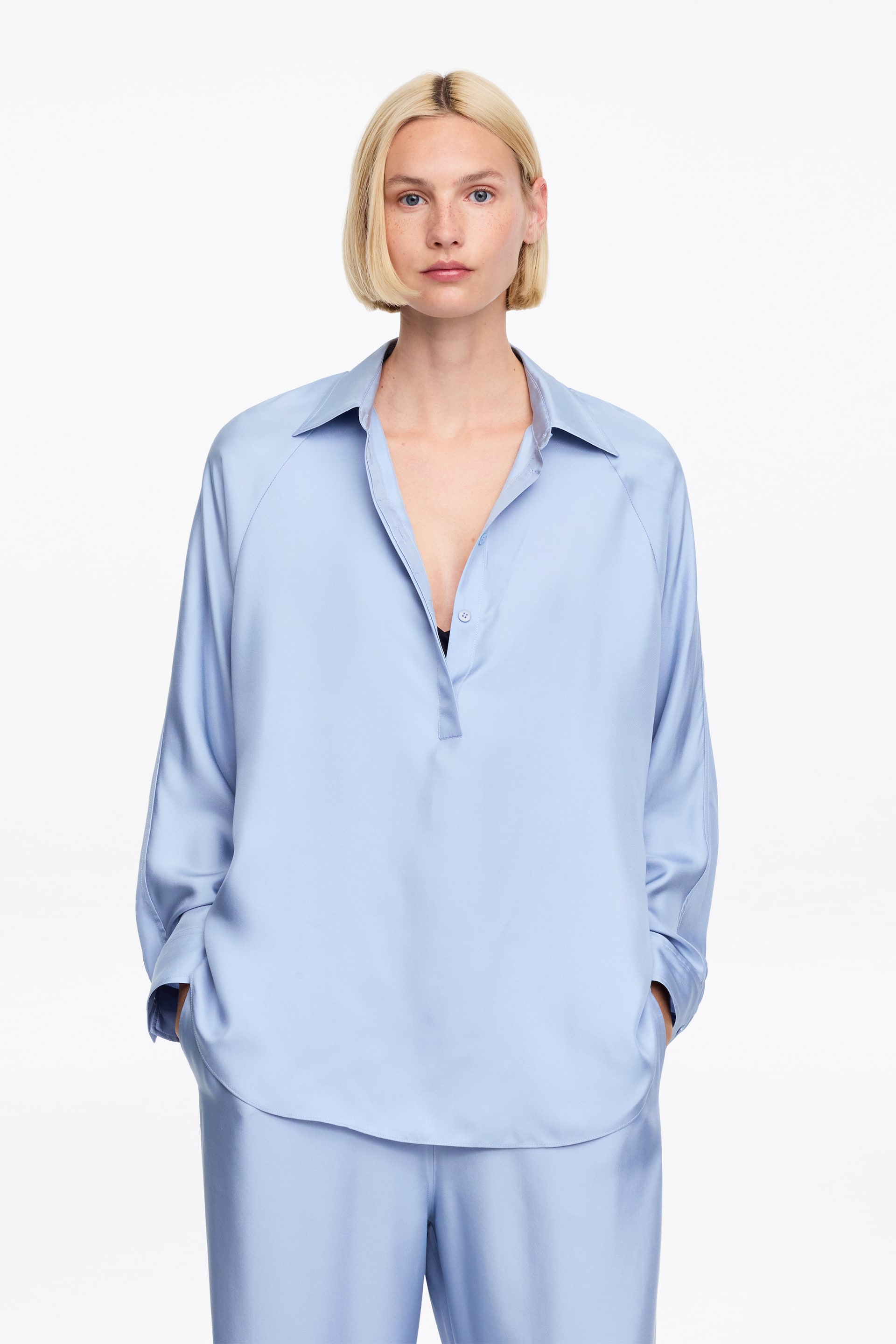 Visualizza immagine più grande: Una camicia fluida azzurro chiaro e pantaloni coordinati dal taglio ampio vengono indossati, rivolti in avanti. La camicia oversize presenta un colletto a punta, un'abbottonatura parziale con un bottone visibile e maniche raglan. Un reggiseno nero è visibile sotto l'abbottonatura aperta.