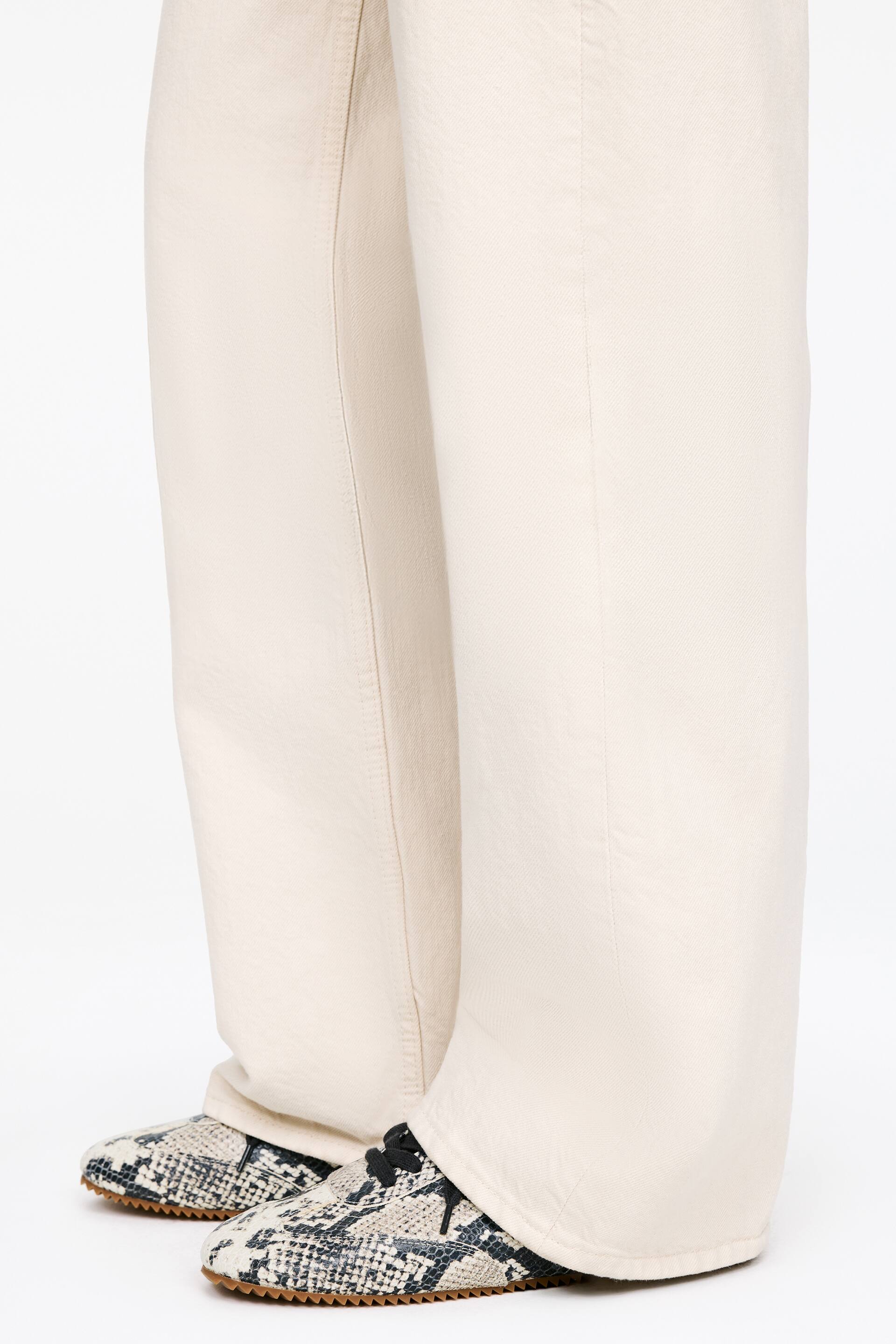 Zobacz większy obraz: Jasnobeżowe, szerokie spodnie o luźnym kroju opadają swobodnie, zakrywając wierzch stóp. Krawędzie spodni spoczywają na butach z wężowym wzorem, które charakteryzują się czarnymi sznurowadłami i brązowymi, ząbkowanymi podeszwami.