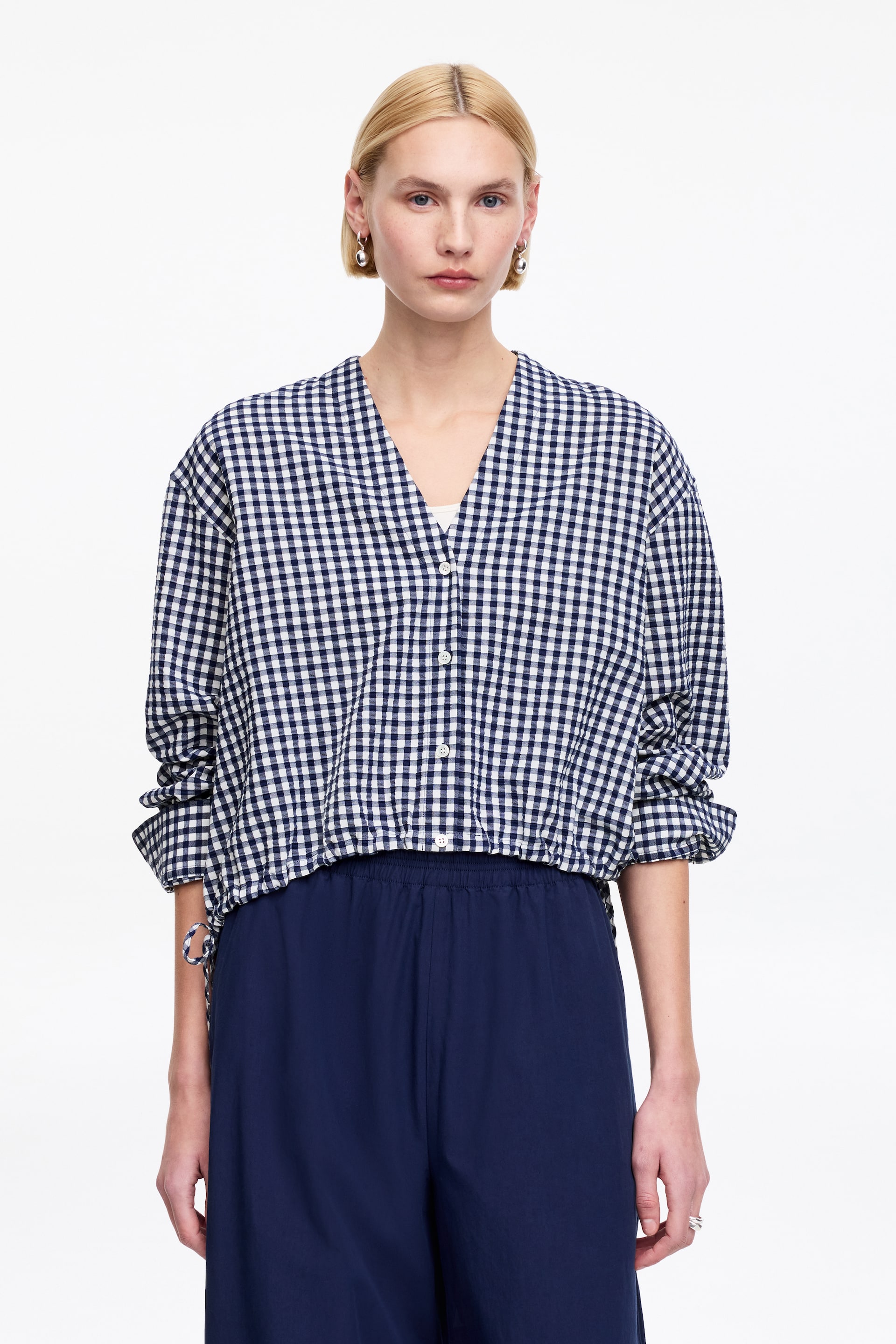 Zobacz większy obraz: Granatowo-biała bluzka w kratkę gingham z dekoltem w serek i zapięciem na guziki, z długimi rękawami podwiniętymi na przedramionach. Bluzka ma krótki, elastyczny dół z widocznym ściągaczem po lewej stronie.