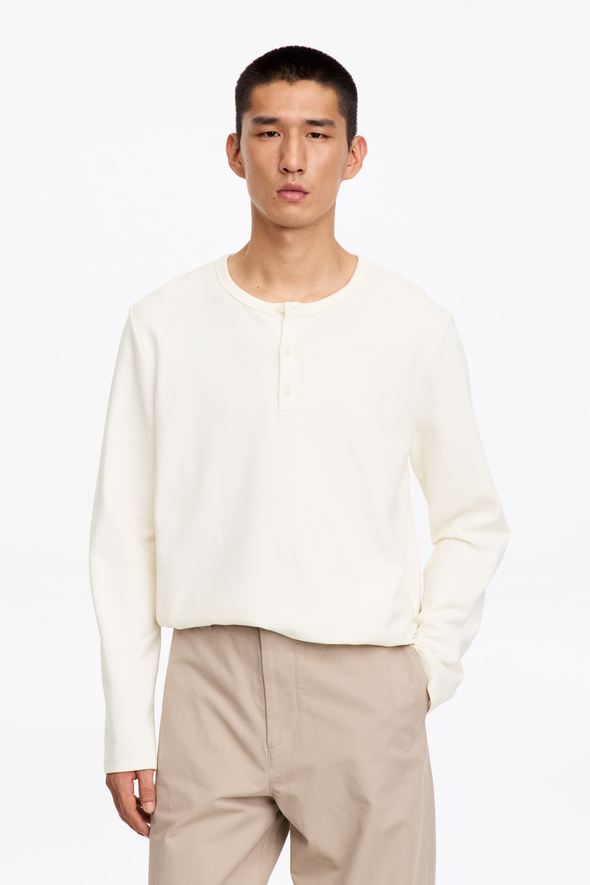 Agrandir l'image: Le mannequin porte une chemise Henley écrue à manches longues, en maille côtelée. La chemise présente un col rond côtelé avec une patte de boutonnage partielle et est rentrée dans un pantalon beige.