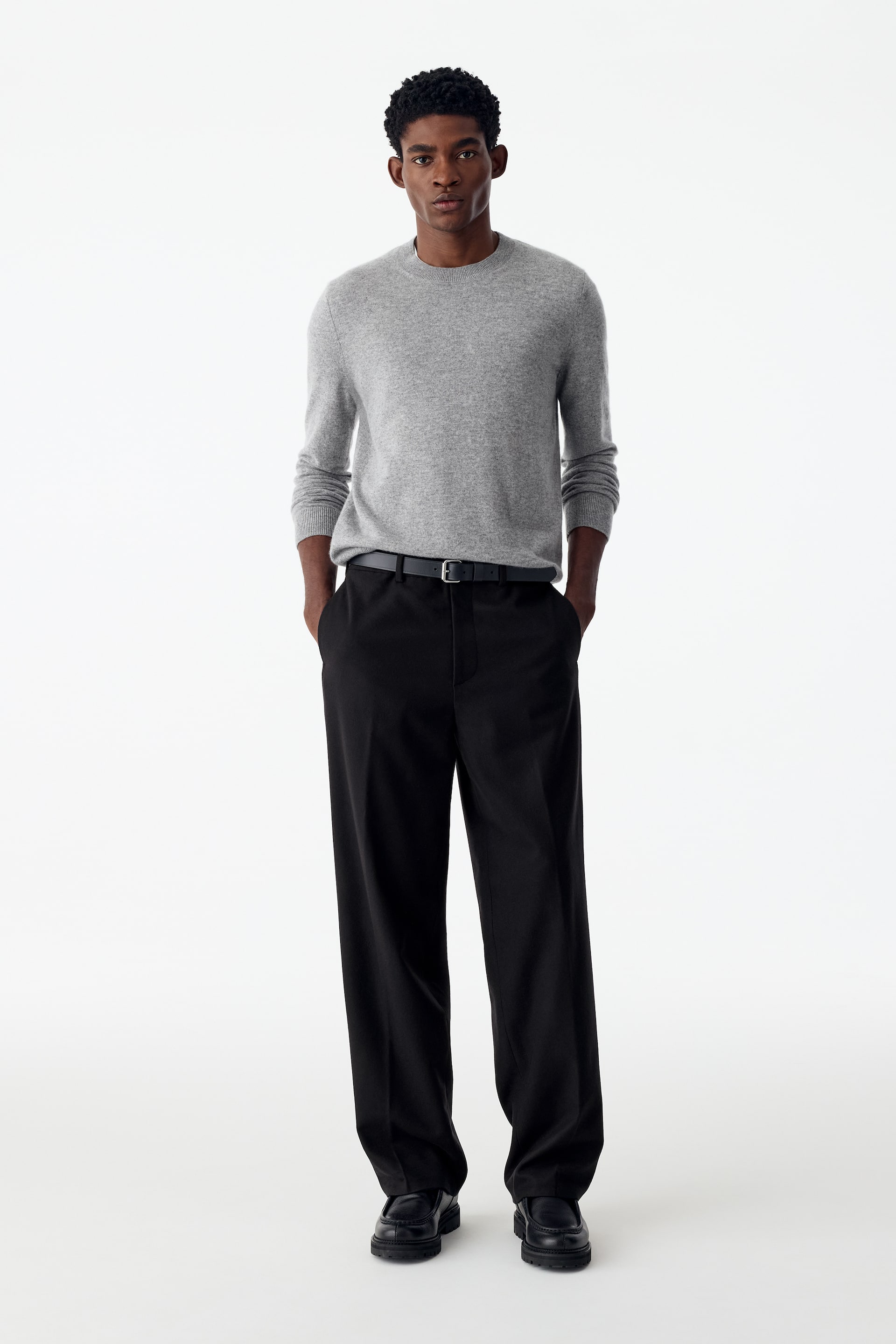 Ver imagen más grande: Estos pantalones negros de pernera ancha con marcadas líneas de planchado los lleva la modelo, que está de pie con las manos metidas en los bolsillos laterales. Los pantalones se combinan con un jersey gris de cuello redondo por dentro, un cinturón negro y zapatos de cuero negros.