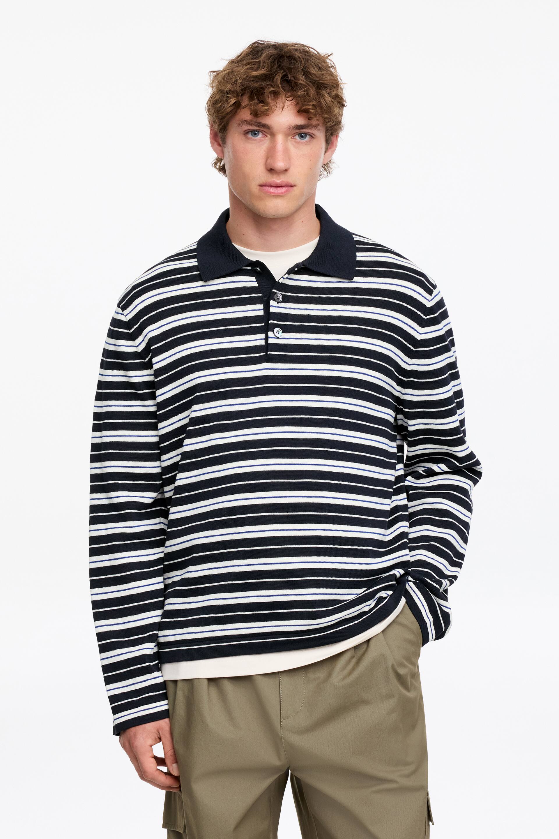 Grotere afbeelding bekijken: Een poloshirt met lange mouwen met zwarte, witte en dunne blauwe horizontale strepen wordt gedragen door het model. Het heeft een donker marineblauwe polokraag en een drieknoopssluiting, en het is gelaagd over een crèmekleurig t-shirt en gestyled met kaki cargobroeken.