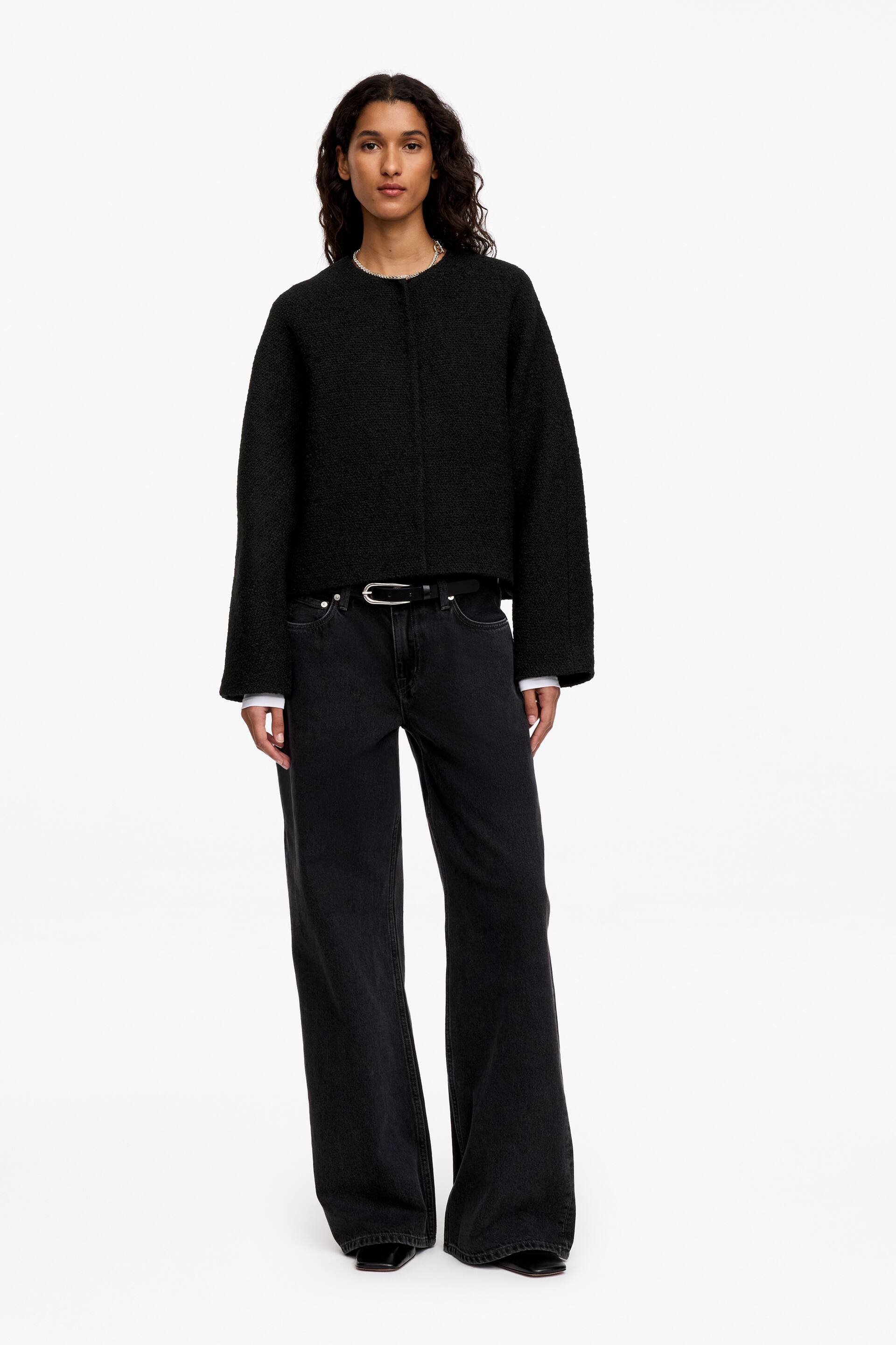 Visualizza immagine più grande: La modella indossa una giacca nera senza colletto realizzata in un tessuto misto lana dalla texture materica. La giacca presenta maniche raglan e una chiusura frontale nascosta, ed è abbinata a jeans neri a gamba larga e a una cintura nera.