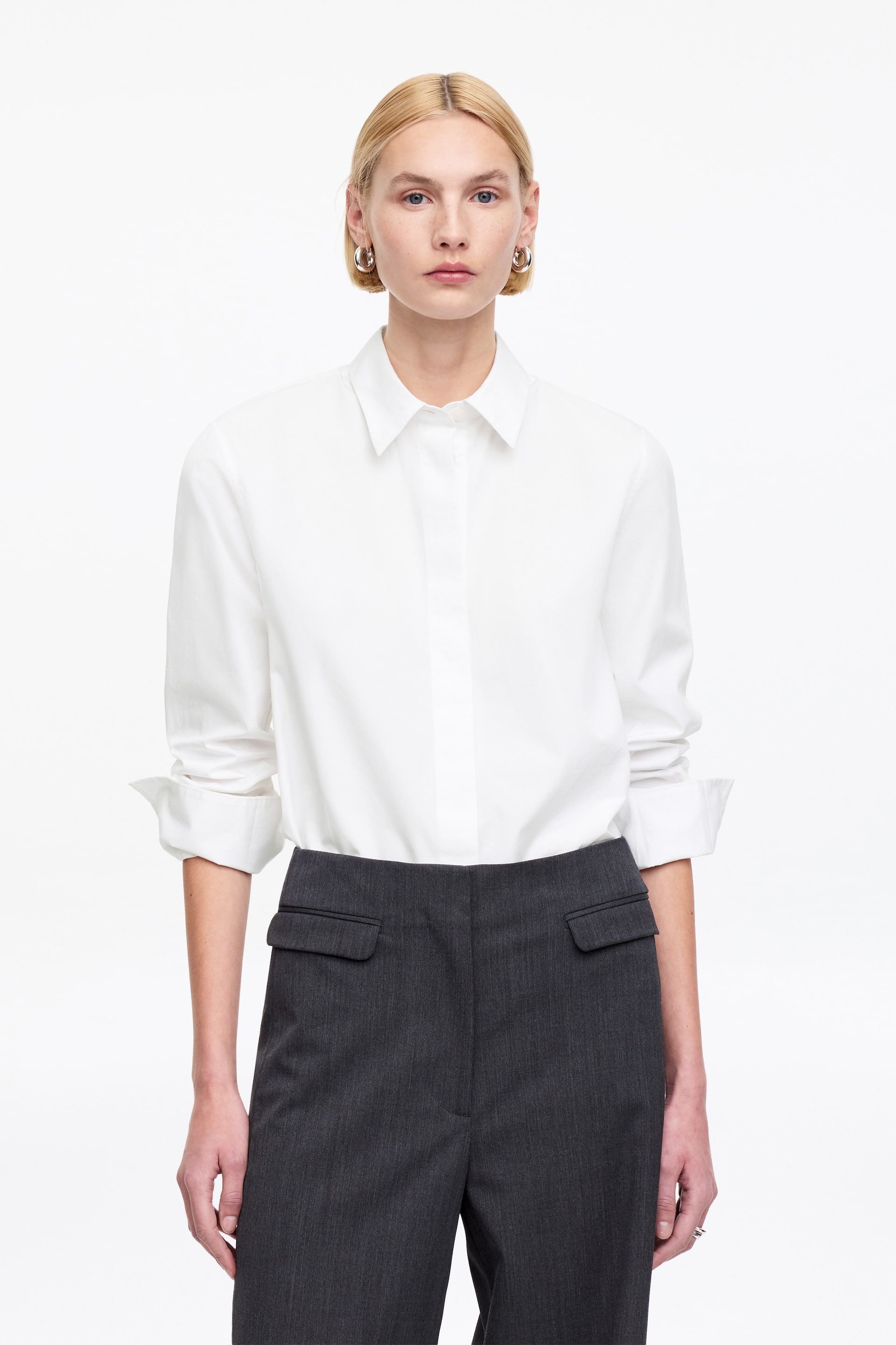 Visualizza immagine più grande: Una camicia bianca con bottoni presenta un colletto a punta e maniche lunghe, arrotolate fino agli avambracci. Questa camicia è infilata in pantaloni grigio scuro, che mostrano due tasche posteriori con patta e una sottile trama verticale.