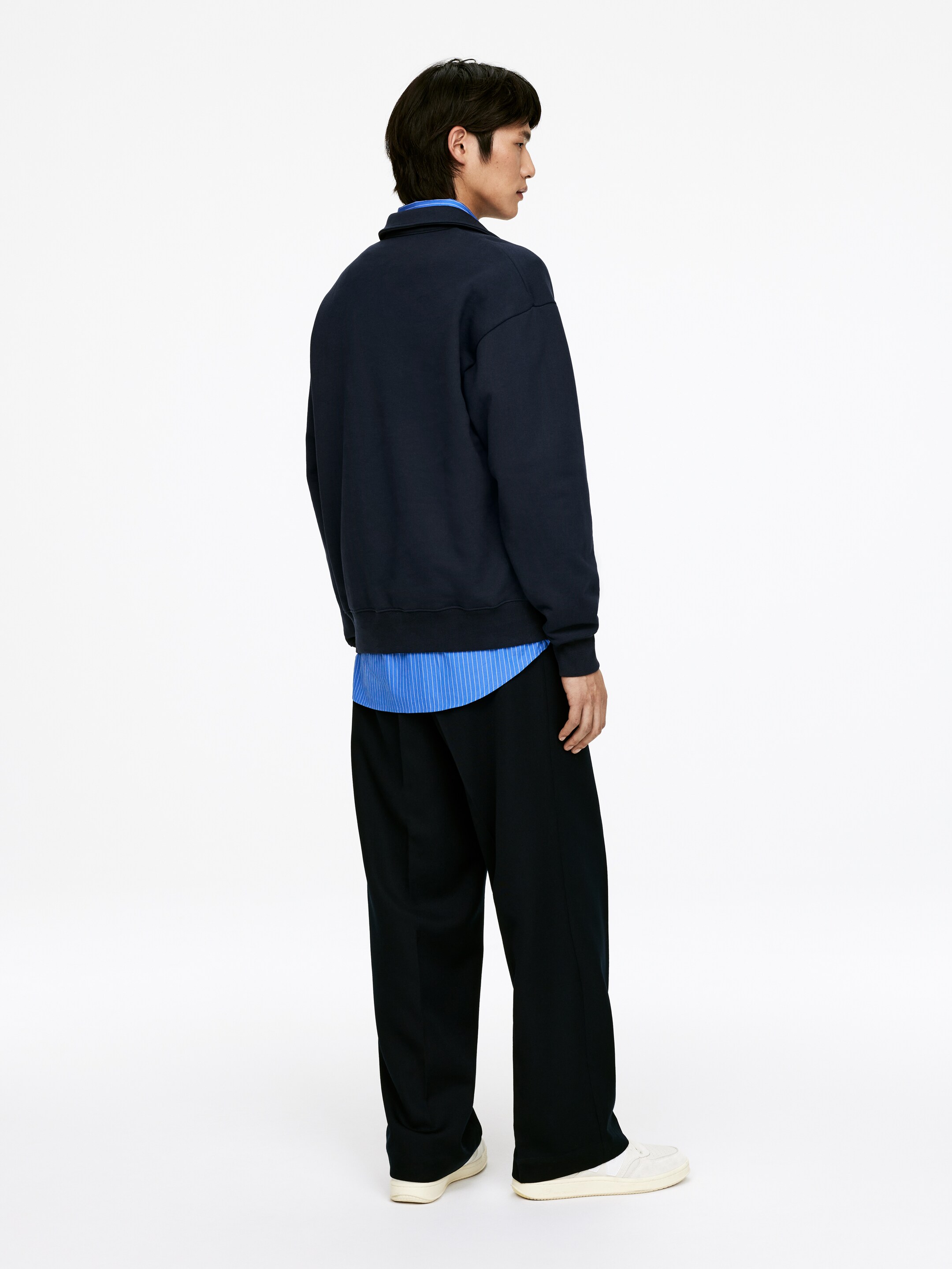 Ver imagem maior: Uma camisola azul-marinho com gola está a ser usada por uma pessoa de costas e ligeiramente virada para a direita, exibindo o seu ajuste relaxado e oversized. Está a ser sobreposta a uma camisa às riscas azul-claro e branca, e está a ser combinada com calças pretas de perna larga e ténis brancos.