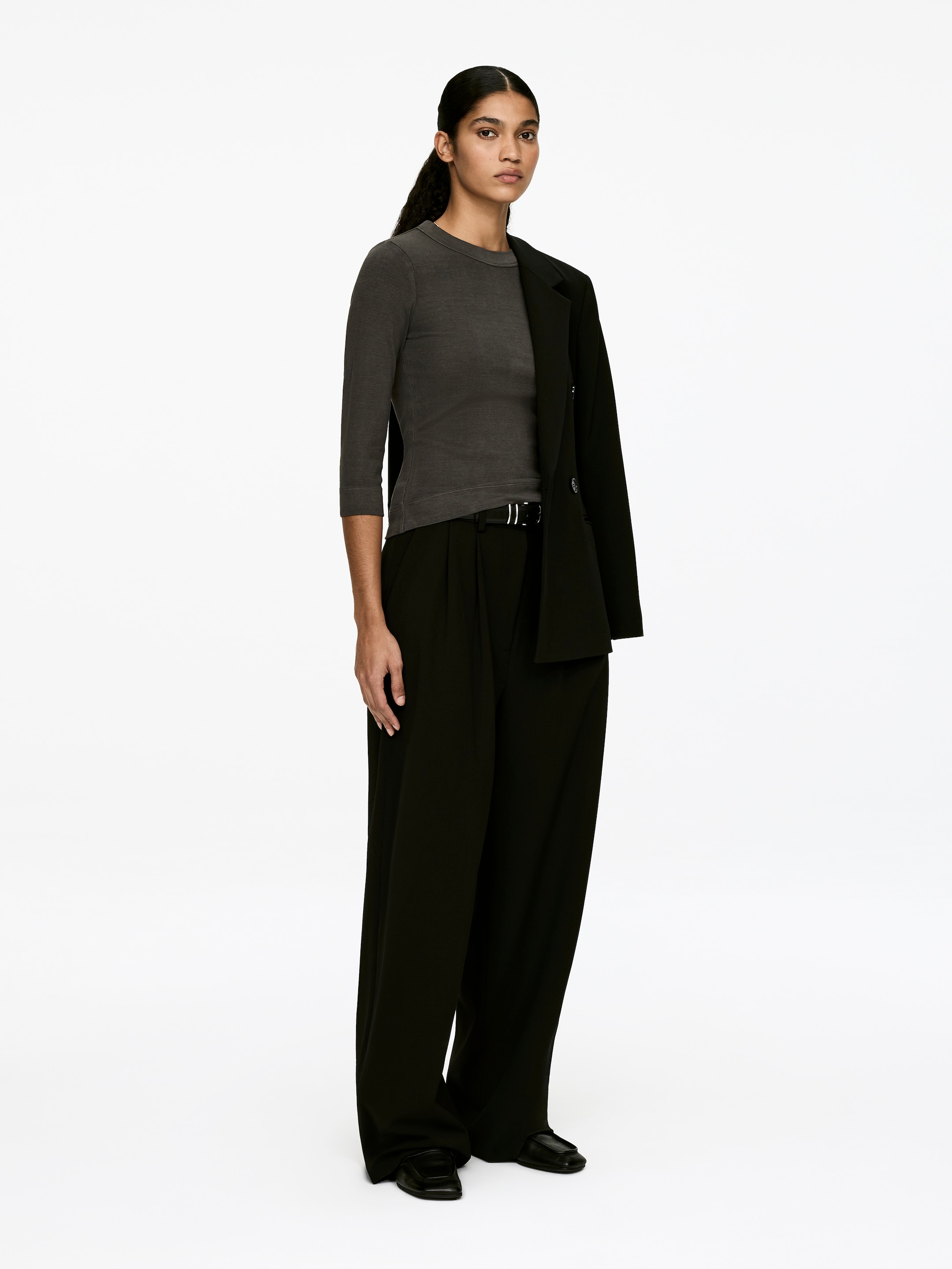 Grotere afbeelding bekijken: Een donkergrijs, slim-fit crew-neck top in geribbelde katoenen jersey, met driekwart mouwen en een zichtbare 3,5 cm bies aan de hals, wordt gedragen door het model. De top wordt gestyled met een zwarte broek en een zwarte blazer die over één schouder is gedrapeerd.
