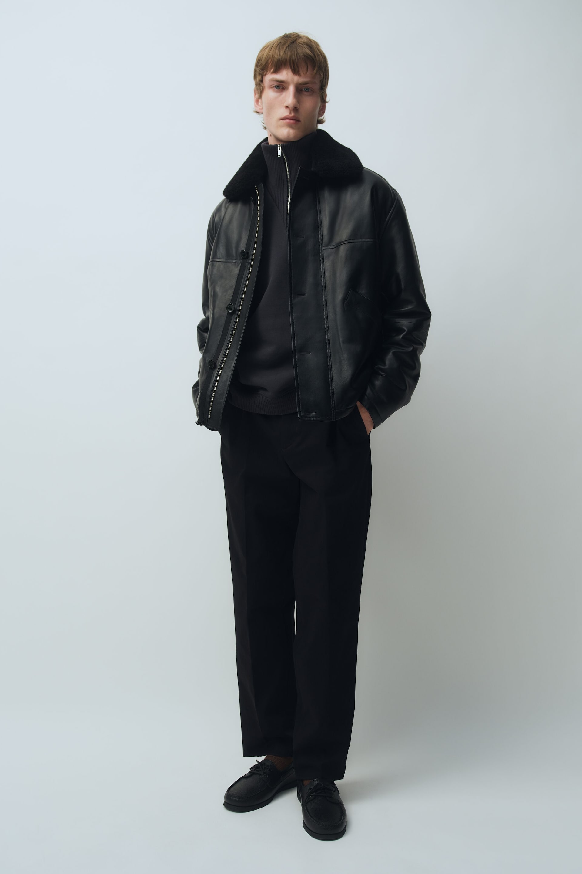 Ver imagem maior: Um casaco de cabedal preto com gola de shearling macia, fecho frontal de correr e pala corta-vento com botões é usado sobre uma camisola de malha preta de gola subida com fecho e umas calças pretas. O casaco é usado aberto, destacando o seu corte descontraído e o design estruturado, numa vista frontal ligeiramente angulada.