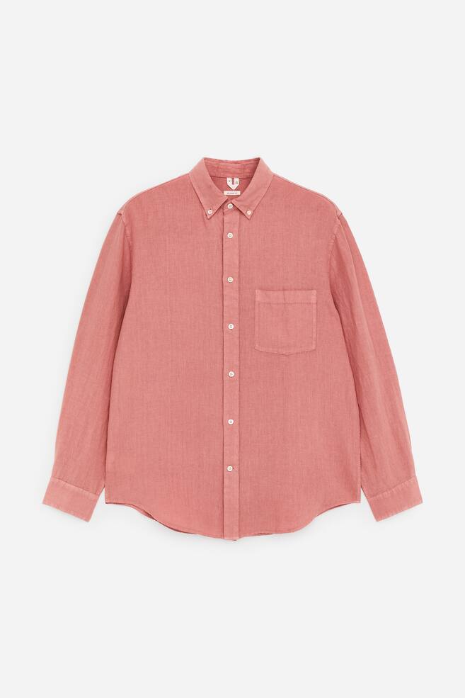 Uma camisa de linho tingida em tom avermelhado-rosado está virada para a frente, exibindo um corte descontraído, uma gola com botões, um bolso único no peito à esquerda, uma bainha curva e punhos com botões. - 1