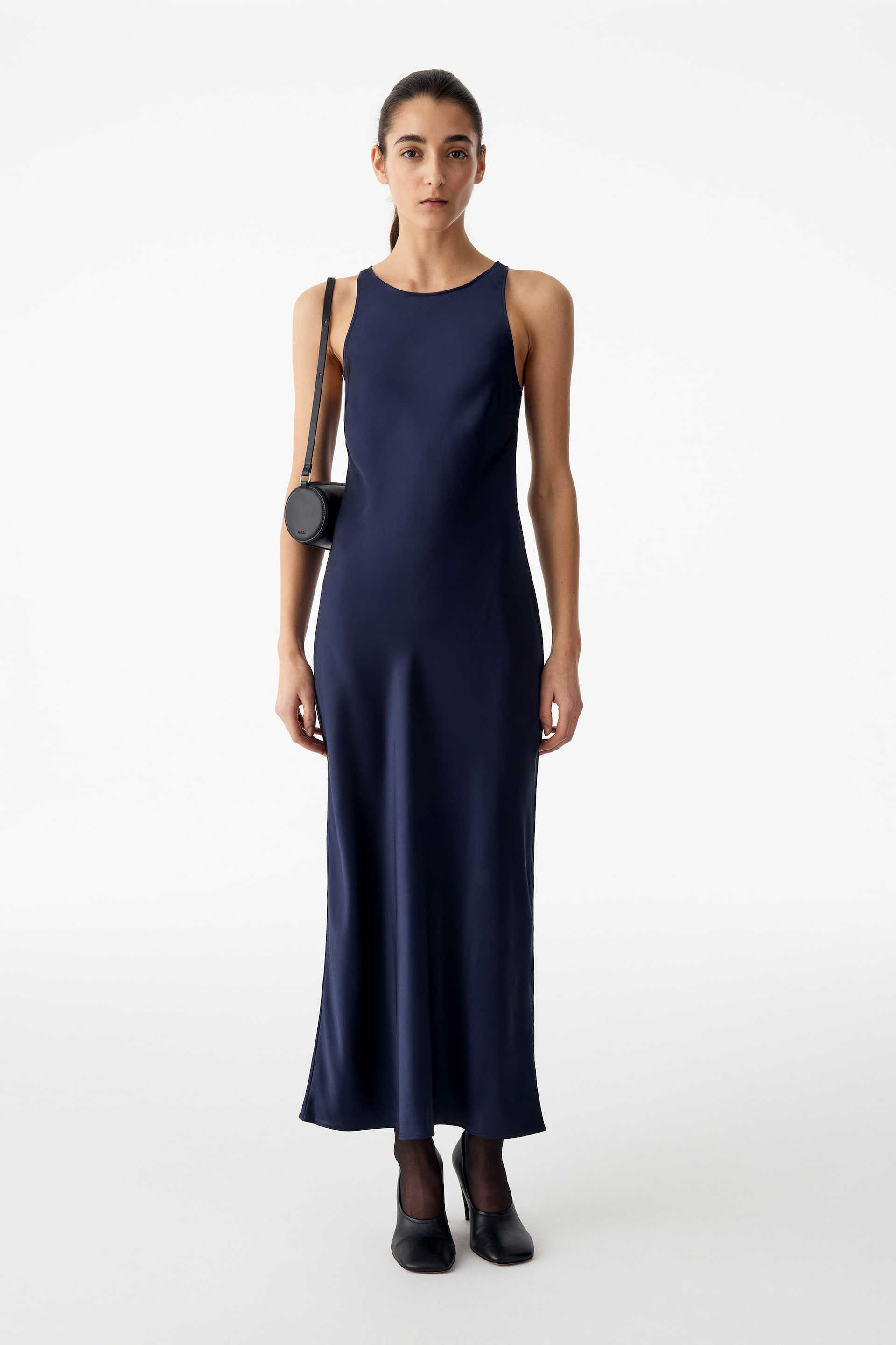 View larger image: A dark navy-blue silk satin slip dress featuring a slim fit, a high round neckline, and a sleeveless, bias-cut midi silhouette is worn by the model. The dress is styled with a small black cylindrical shoulder bag, sheer black tights, and black court shoes.