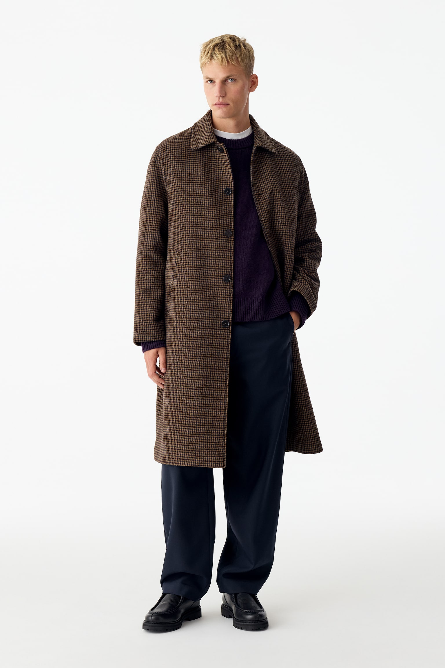 Un long manteau à simple boutonnage arborant un motif pied-de-poule brun et noir est présenté de face, mettant en valeur un col à pointes classique et une fermeture à cinq boutons. Le manteau présente des manches longues et deux poches passepoilées obliques sur les côtés. - 1